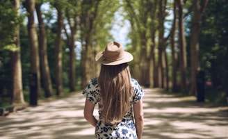 Woman with hat walks down a tree-lined path.