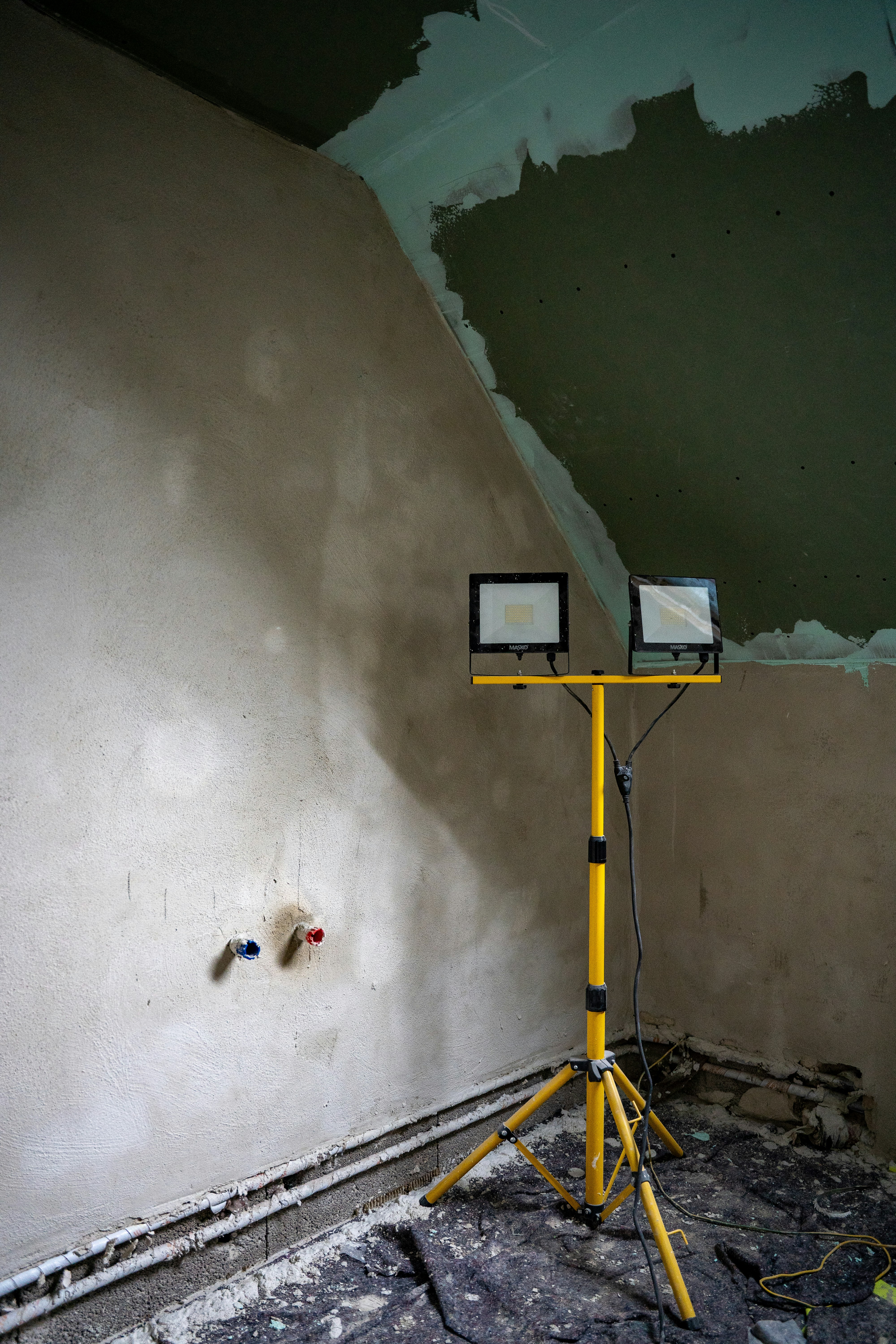 A raw, unfinished corner of a room under renovation, illuminated by a bright dual-head construction lamp. Exposed plumbing and patchy drywall reveal the gritty backbone of a transformation in progress.