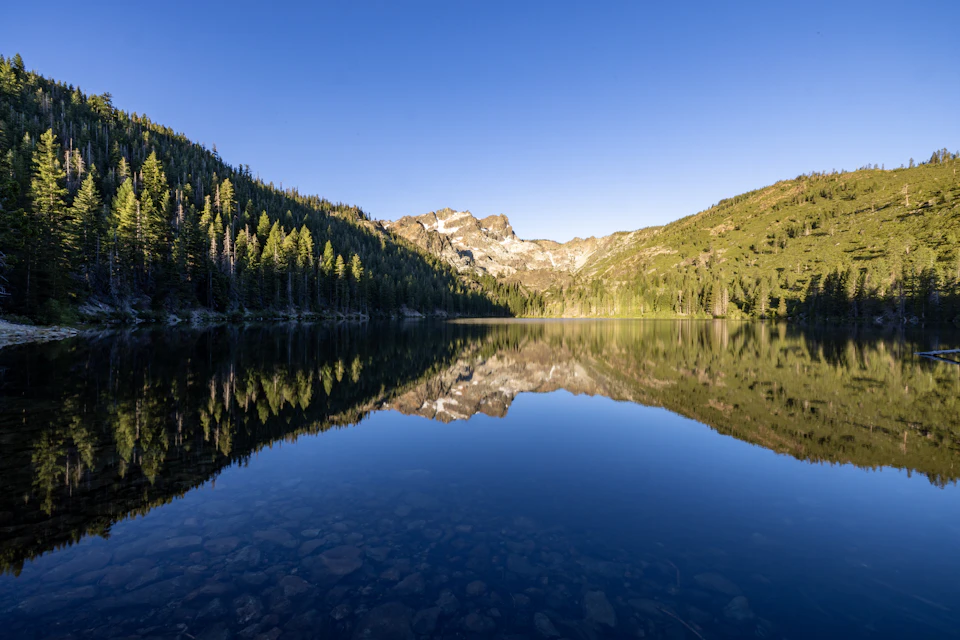 The image depicts a serene lake nestled within a mountainous landscape. The calm, deep blue water reflects the surrounding trees and mountains, creating a mirror-like effect that enhances the tranquility of the scene. The perspective is from the shoreline, looking out towards the mountain range in the background. The sky above is a clear blue, suggesting a bright and sunny day.