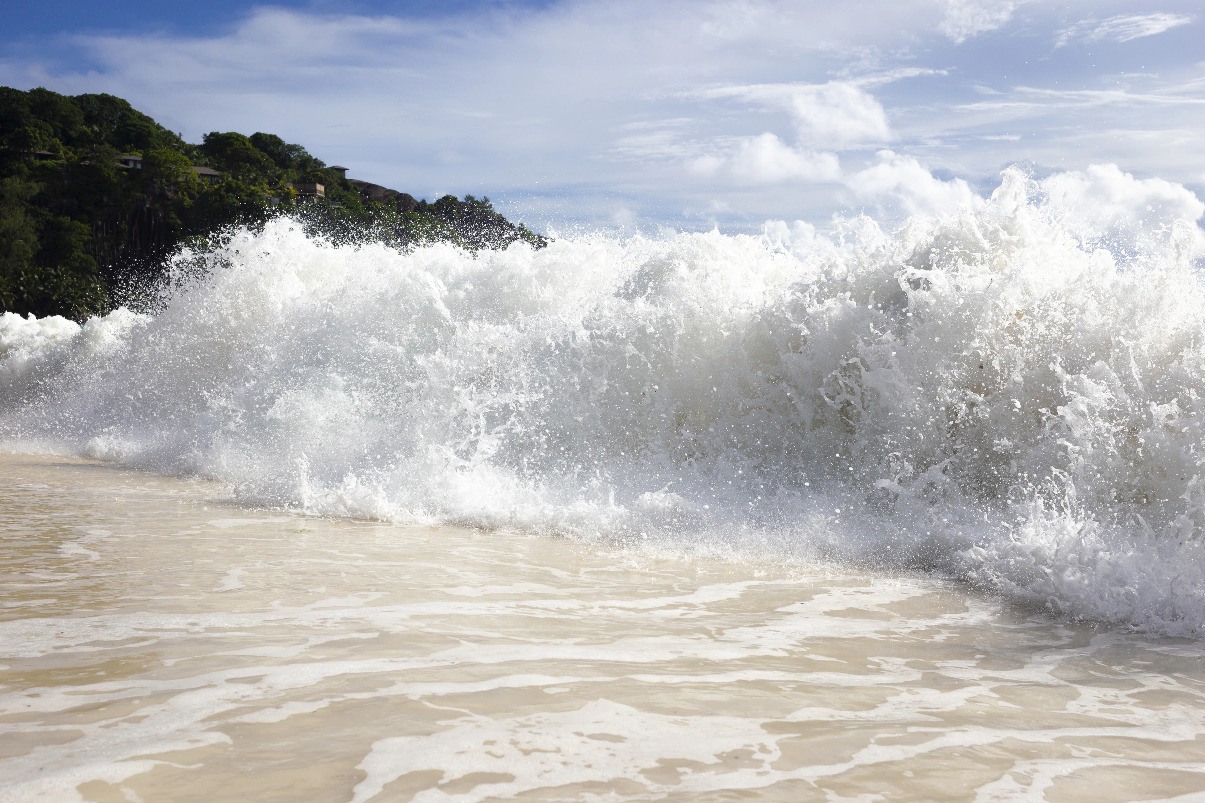 A frothy turquoise wave breaks onto white sand backed by a green forested hillside.