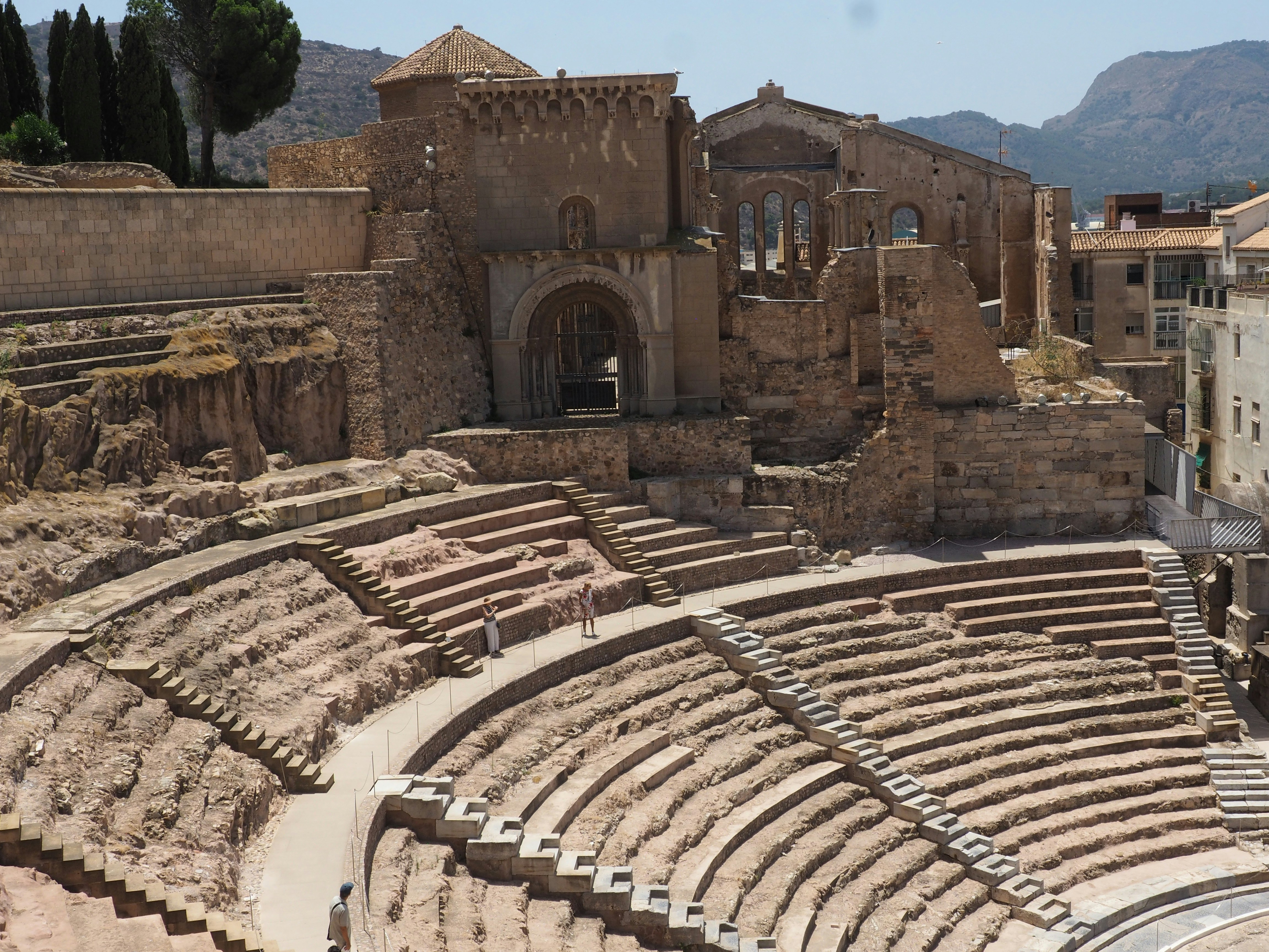 Cartagena - An expansive view of the magnificent Roman Theatre in Cartagena, Spain, bathed in natural light. The ancient tiers of seating are clearly visible, leading the eye down towards the stage area. The historical built structures stand behind the seating. This image truly conveys the grandeur and scale of this remarkably preserved archaeological site, The people look so small. It offers a compelling glimpse into the city's rich past.