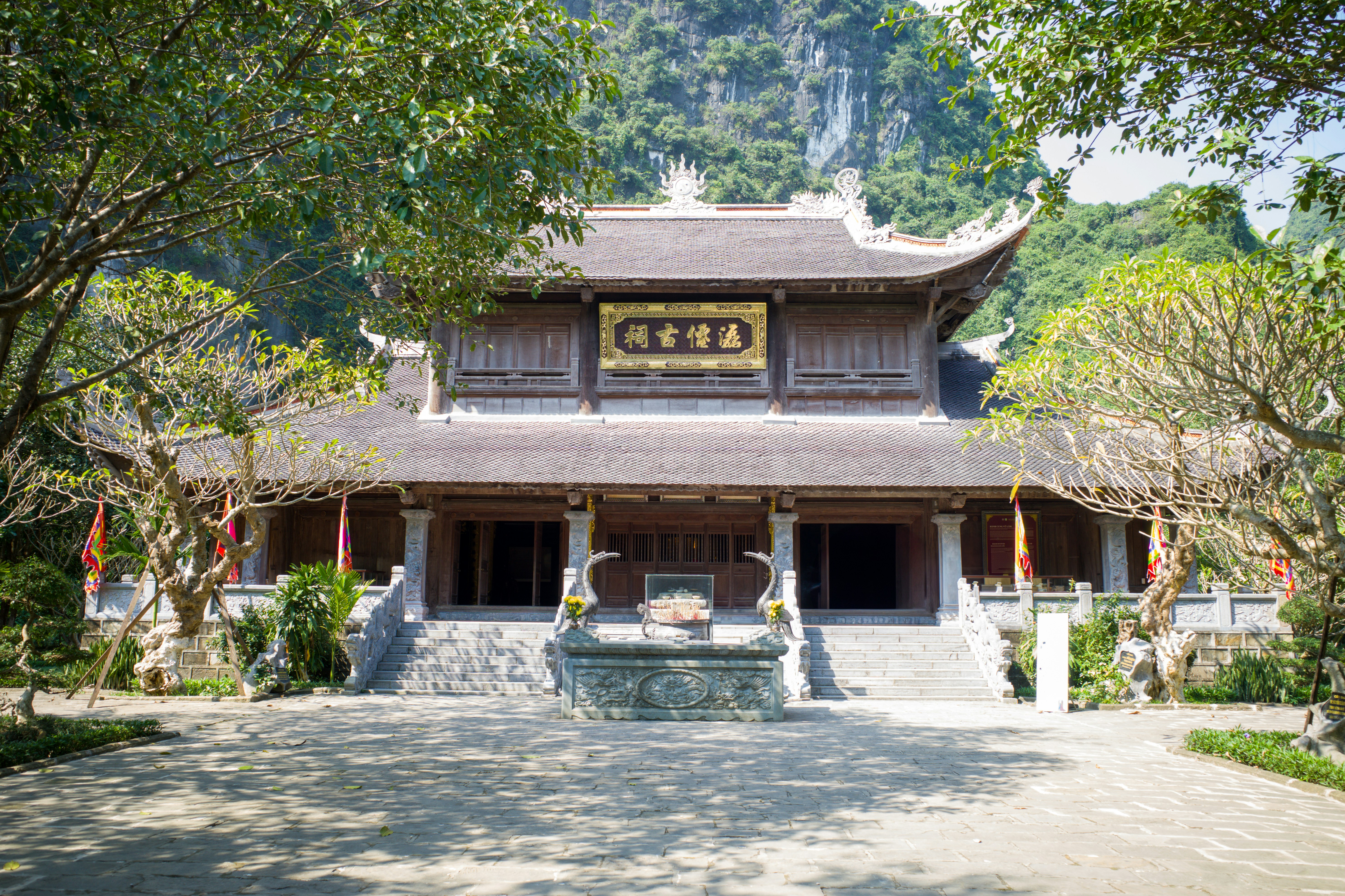 A traditional wooden temple with ornate roof carvings and a stone incense burner stands among trees. The courtyard is quiet, framed by trimmed plants and mountain cliffs behind, representing serene temple architecture typical of East or Southeast Asia