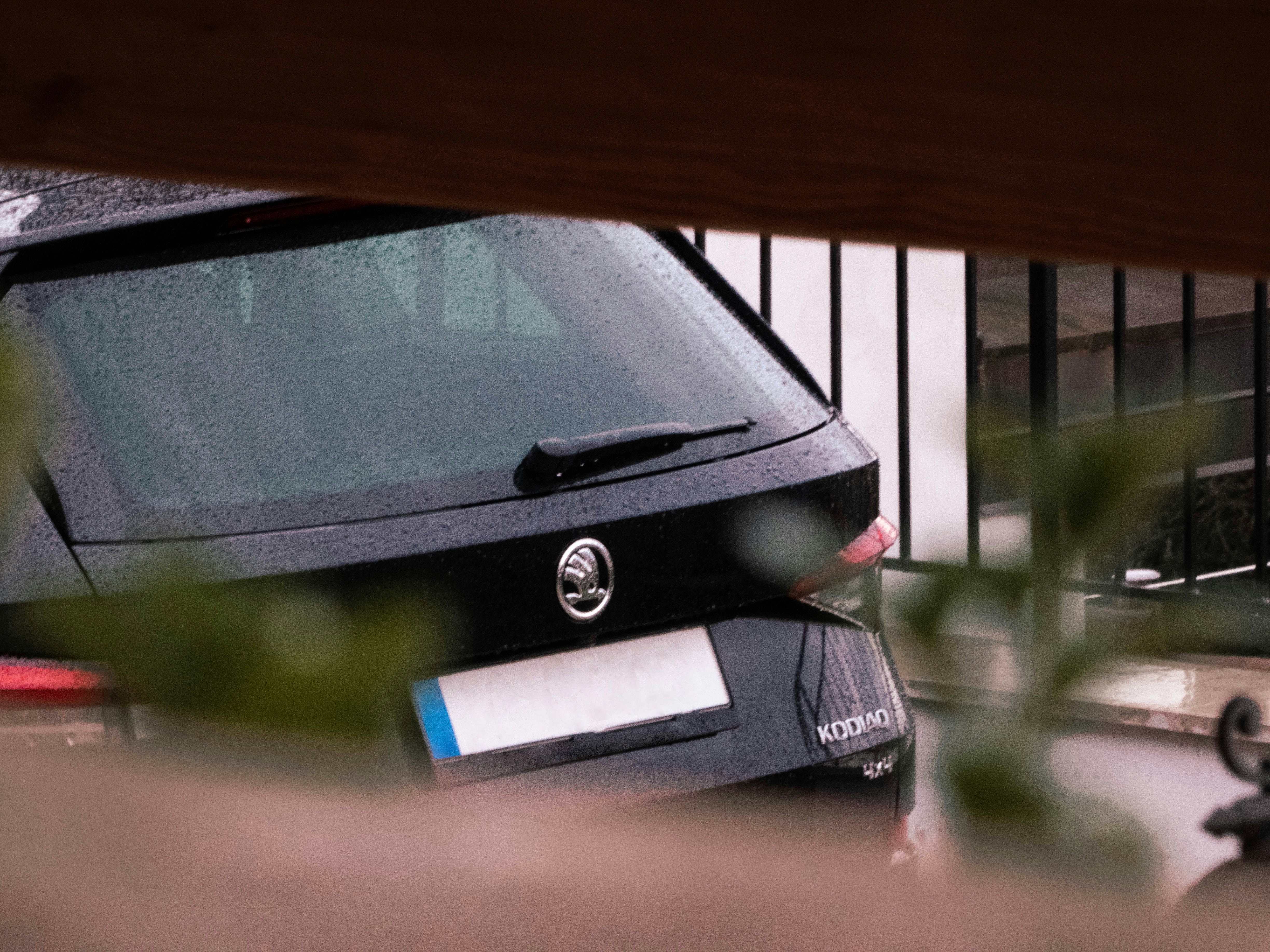 A black Škoda Kodiaq 4x4 is parked behind a fence on a rainy day. The rear of the vehicle is visible, showing the brand emblem, rear windshield wiper, and part of the license plate, which has been obscured. Water droplets are present on the car's surface and window, and some foliage is partially covering the view.