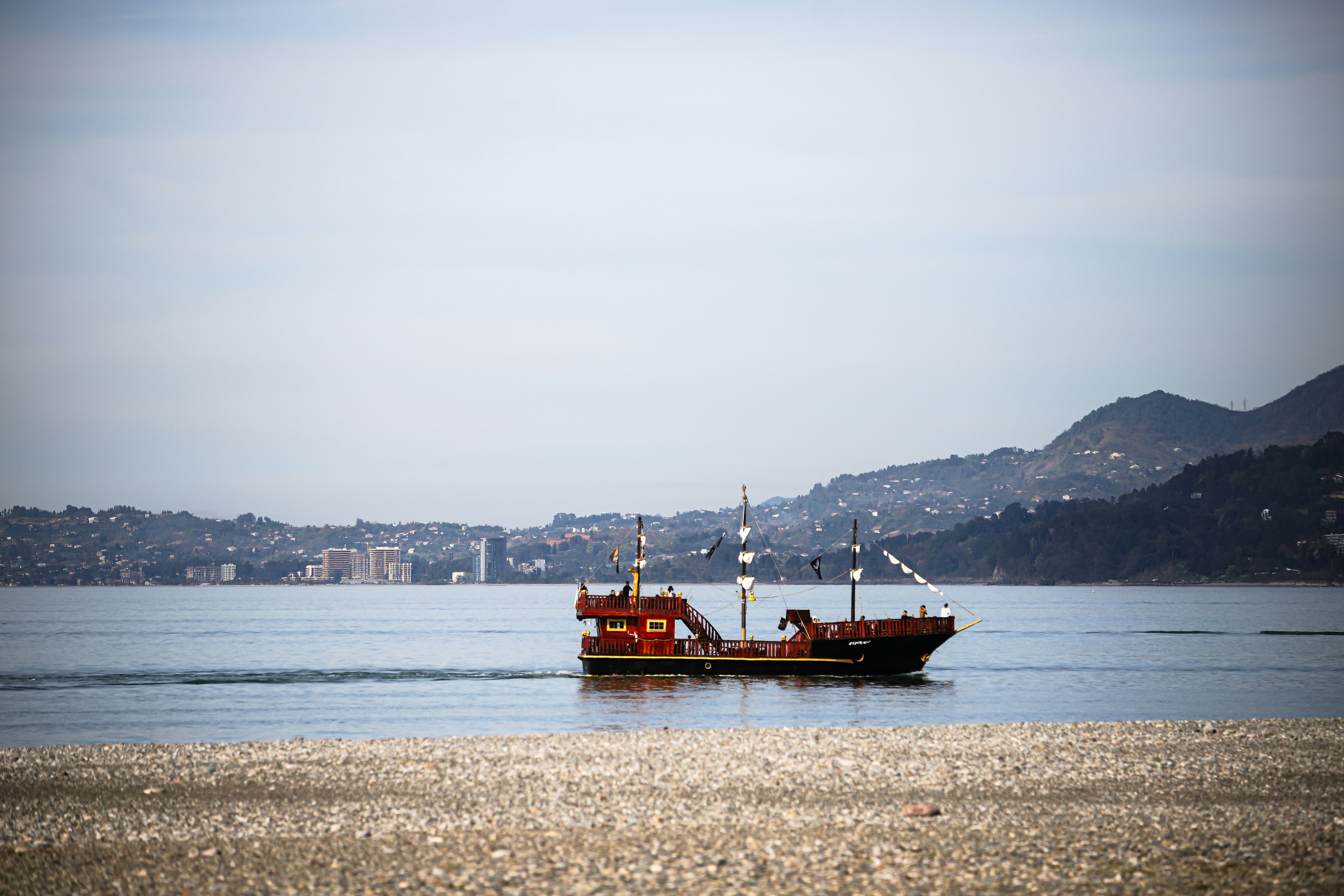 A classic-style wooden ship sails gracefully near the pebble shores of Batumi, Georgia. With its dual masts and black flags, it evokes maritime history against a backdrop of modern buildings and lush hills—a timeless blend of tradition and urban charm.