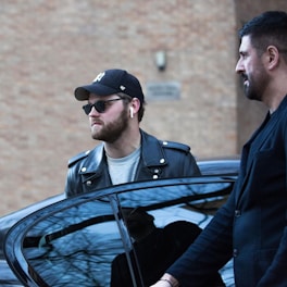 Man in leather jacket exits car with security