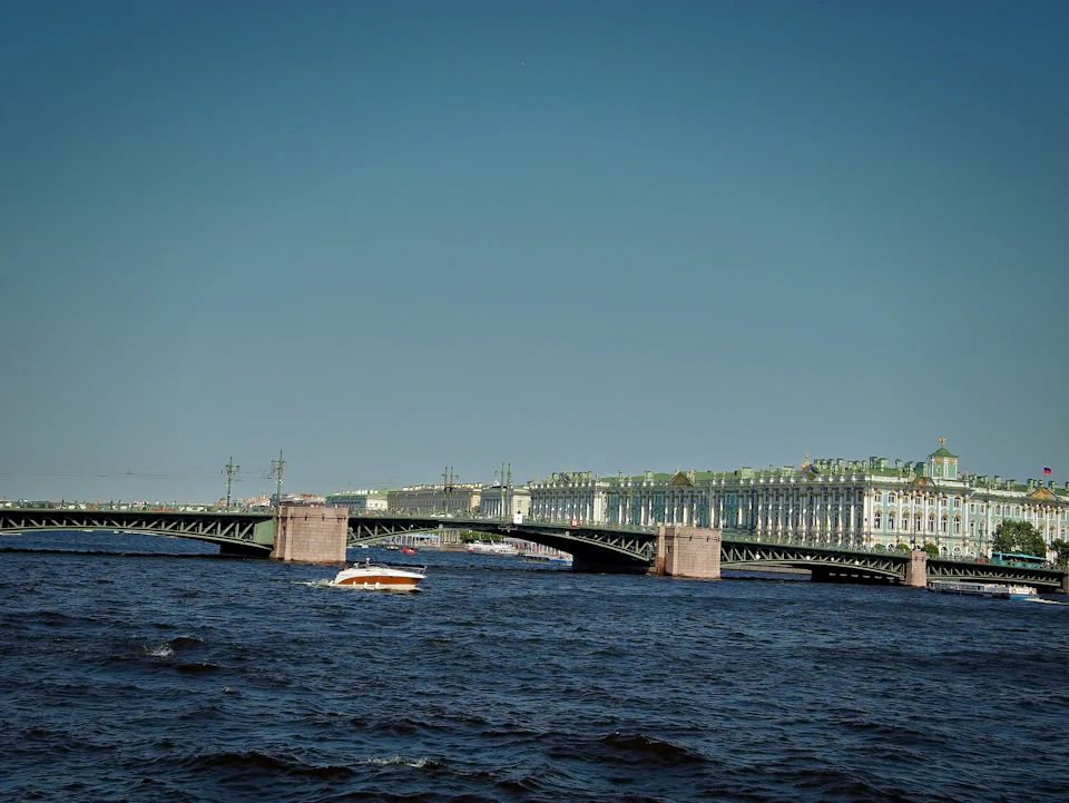 The image depicts a serene scene on the Neva River, with a large green bridge spanning across it. The bridge is adorned with two towers and features multiple arches that add to its architectural charm. On the river below, there are several boats of varying sizes scattered throughout the water, including an orange boat in the foreground and another one further away.

In the background, a majestic building can be seen, possibly a palace or a large residence. The sky above is a clear blue, providing a beautiful backdrop to this picturesque scene.