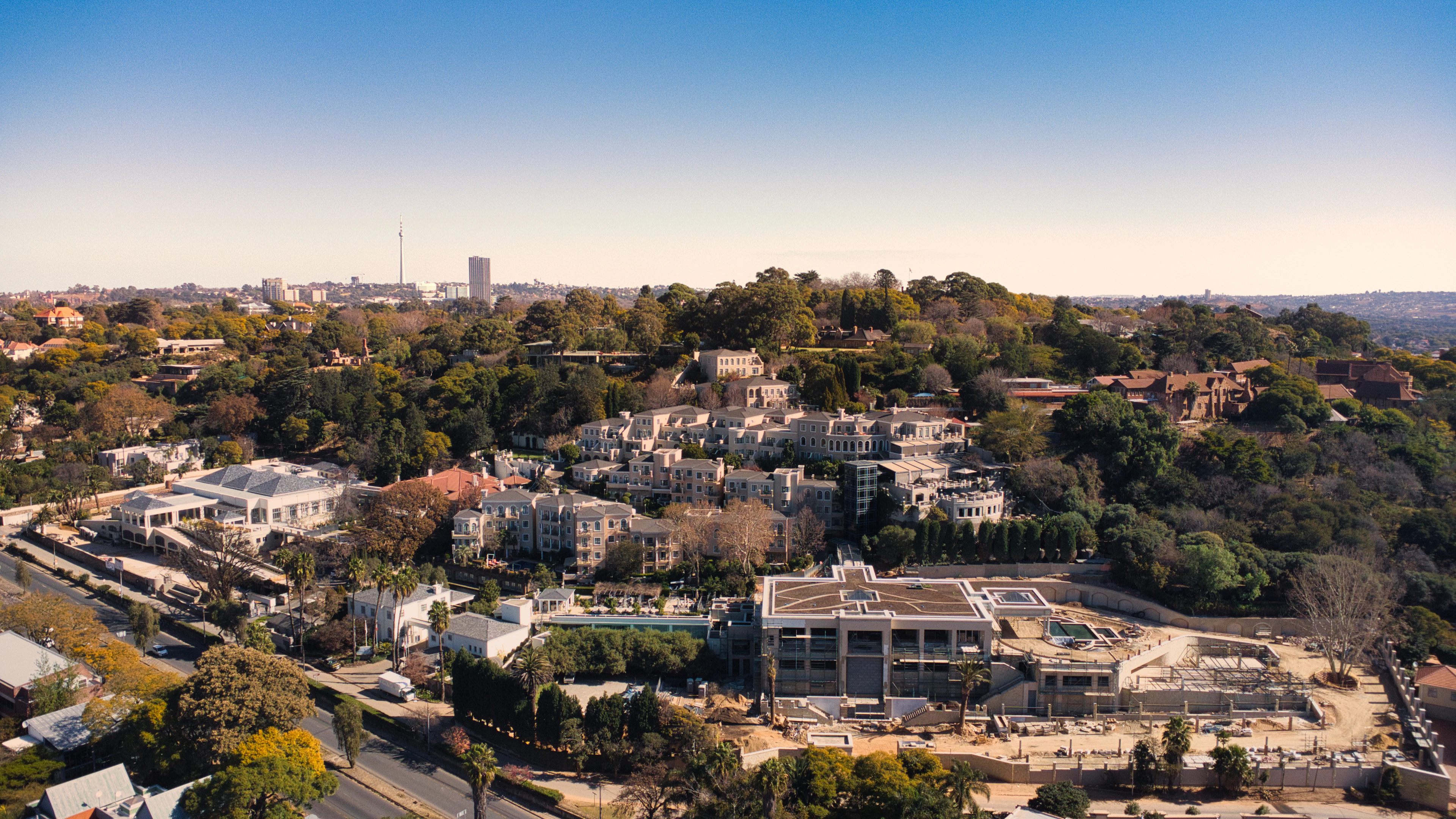 The wooded Westcliff ridge in Johannesburg from the air: white-walled mansions and stepped hotel terraces threaded into the trees along the slope, with the northern suburbs, the Hillbrow Tower and the distant city skyline on the horizon in late-afternoon light.