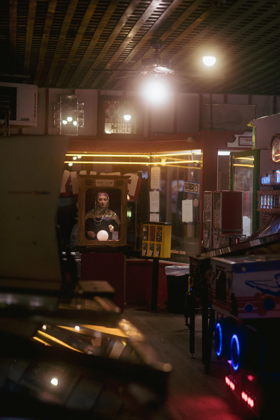 The image depicts a dimly lit room with various arcade games and equipment, including a large claw machine on the right side. A person is visible in the foreground, possibly playing one of the games or observing the surroundings. The room has a wooden floor that adds to its rustic charm, and the ceiling features exposed beams, creating an atmosphere reminiscent of a vintage arcade.