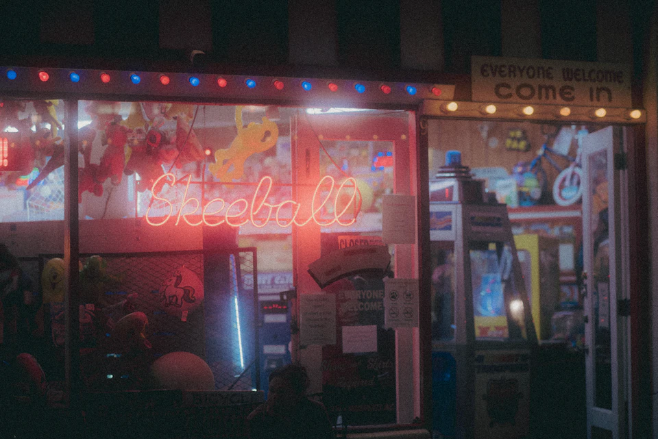 The image shows a neon-lit storefront with a "SKEWBALL" sign, indicating that it is an arcade or gaming establishment. The store's name is prominently displayed on a window, and there are several other signs visible throughout the scene. A person can be seen sitting inside the store, likely enjoying some of the games available for play.

The exterior of the building features a striped awning, adding to its vibrant atmosphere. In front of the store, there is a display with various toys, suggesting that it caters to children and families who visit the arcade or gaming establishment.
