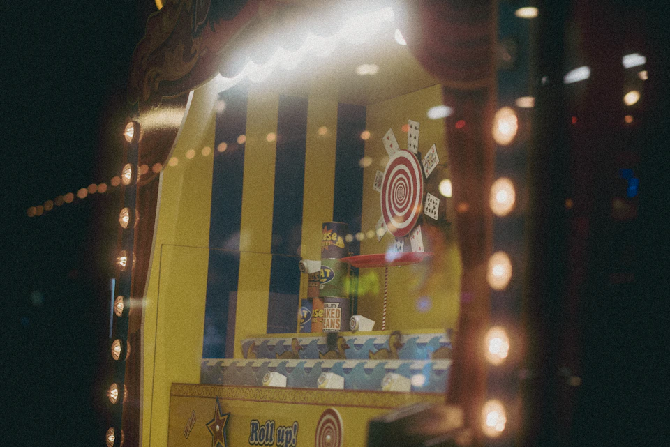 The image shows a carnival game booth with a yellow and blue striped tent, illuminated by white lights that cast a warm glow on the scene. The booth features a target board with red targets and a scoreboard displaying "ROLLING DICE". A small figure of a man is also present in the booth, likely representing the game's operator or mascot.

The perspective of the image is taken from inside the booth, looking out onto the street outside. This creates an interesting contrast between the vibrant colors of the carnival booth and the dark background, making it appear as if the viewer is part of the scene.