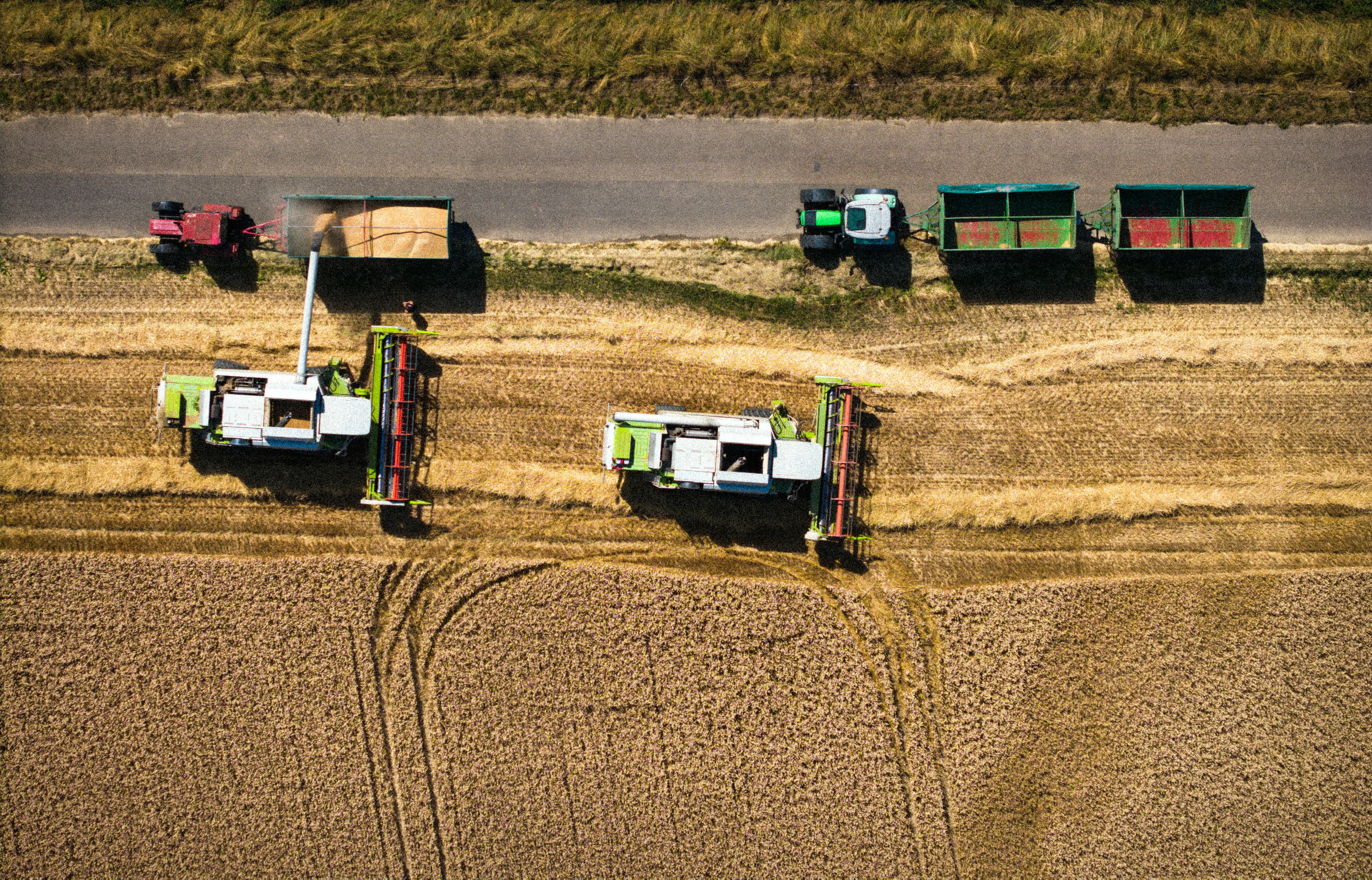 Aerial views of two combine harvesters operating in a wheat field, transferring harvested grain into trailers pulled by tractors parked along an adjacent rural road