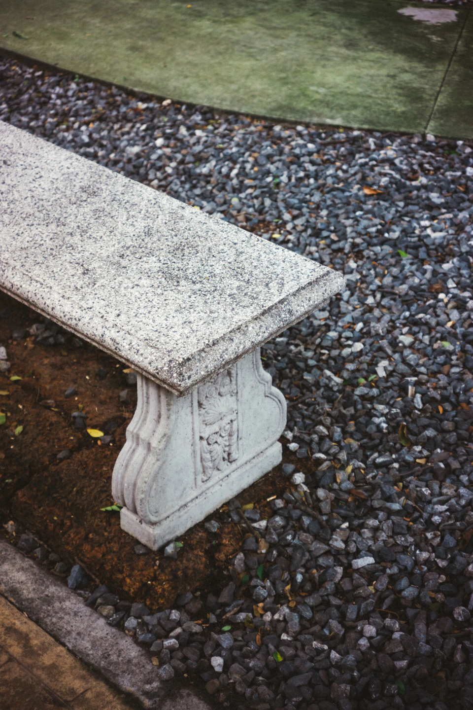The image shows a white stone bench with a curved backrest and armrests, situated on a gravel path surrounded by small rocks. The bench is positioned against a backdrop of green grass and trees, creating a serene outdoor setting.