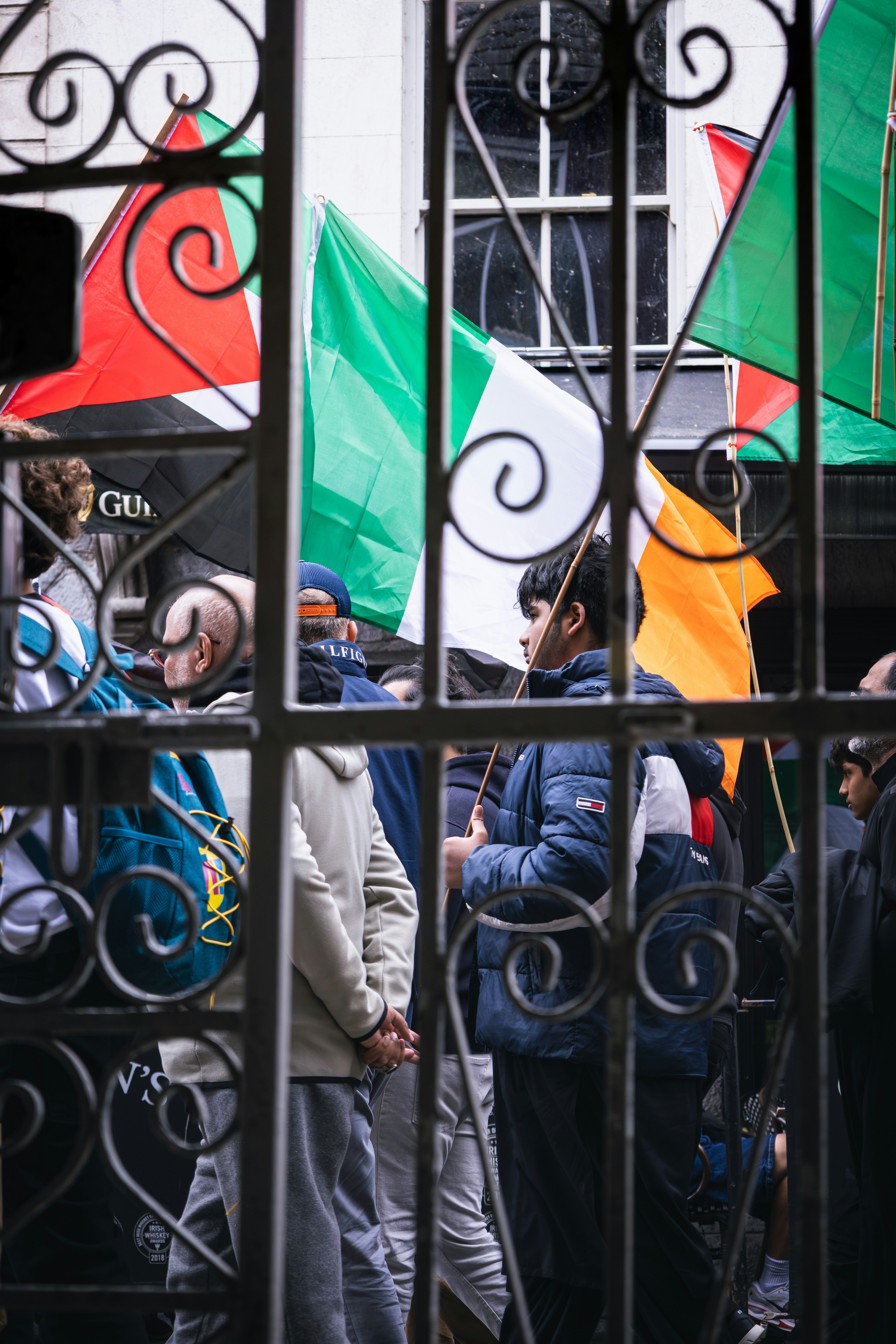 People gather behind a wrought iron gate with flags.