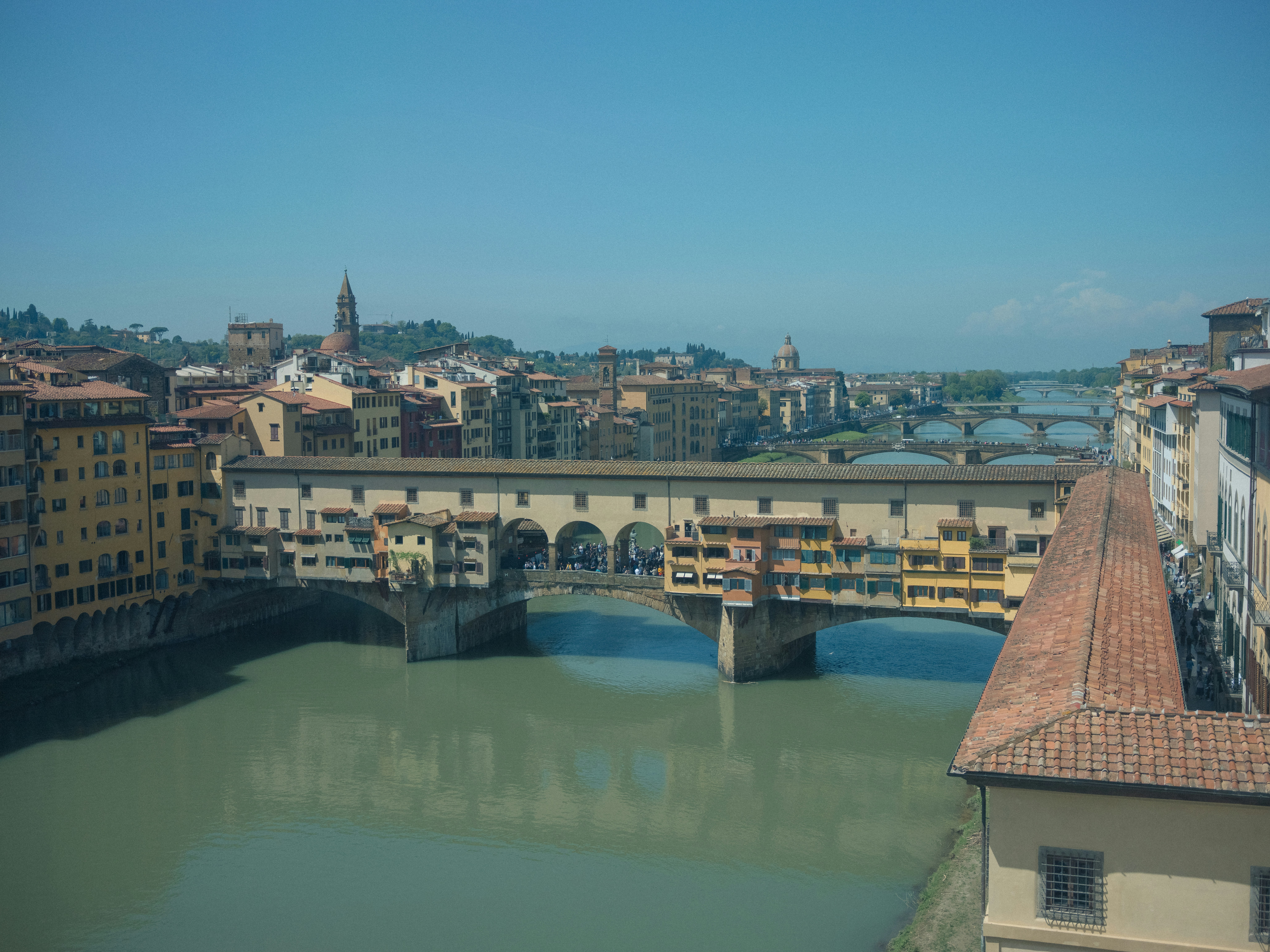 Veduta soleggiata diurna di Ponte Vecchio a Firenze, Italia, con i colorati edifici lungo il fiume e l'Arno che scorre sotto.