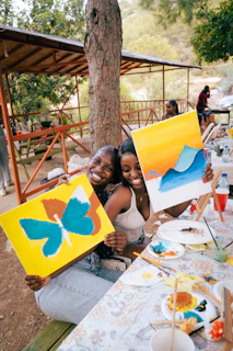 Two women smile while holding their paintings.