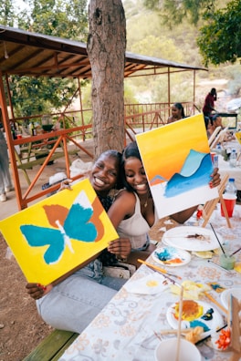 Two women smile while holding their paintings.