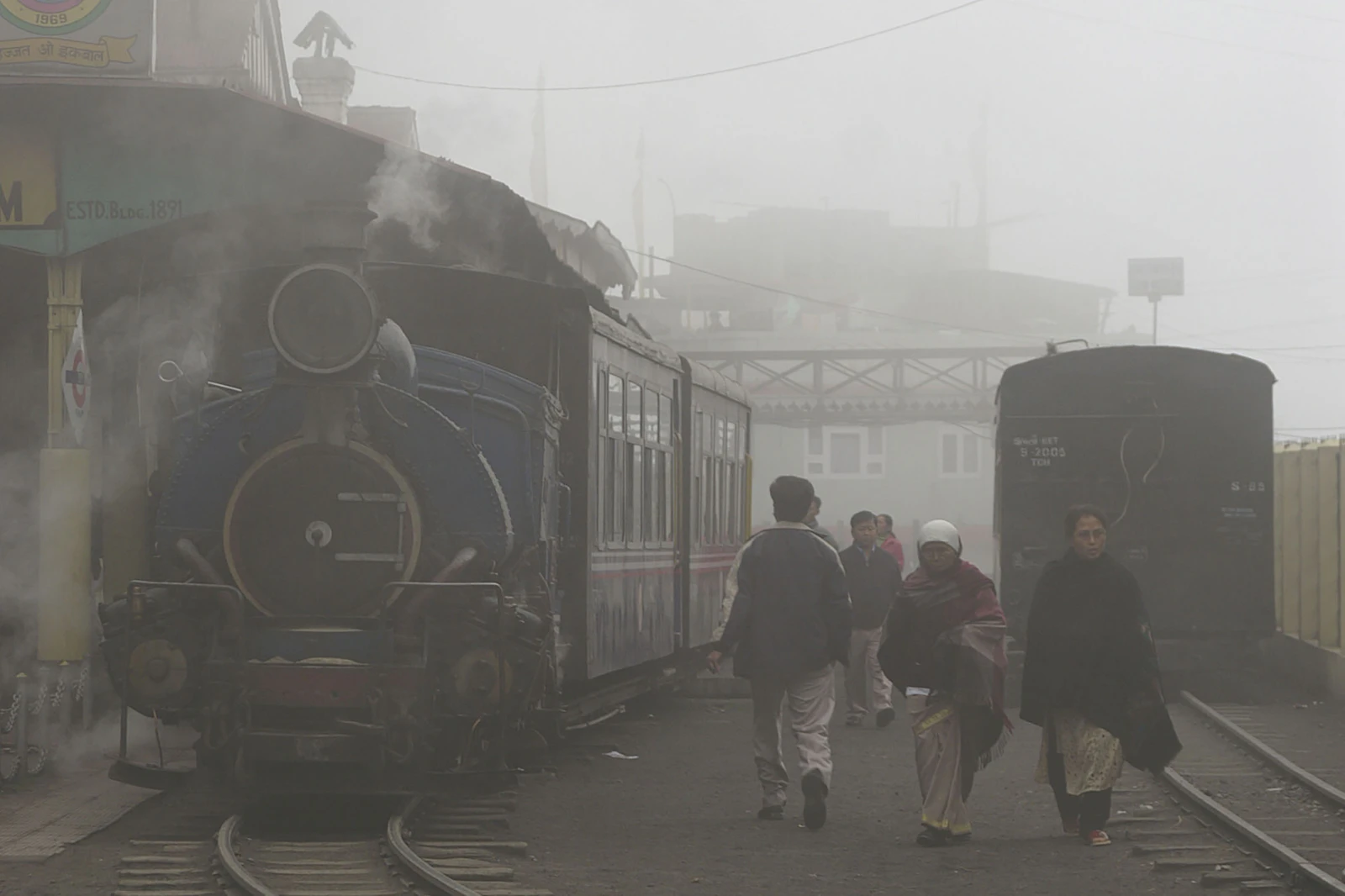 A blue steam engine of the toy train breathing smoke at a foggy Darjeeling station as travellers pass