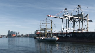 A tall ship sails near cargo cranes in a harbor.