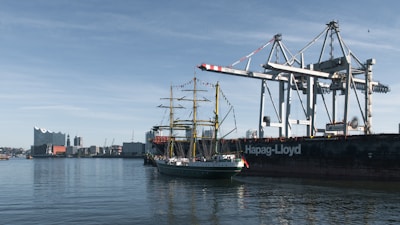 A tall ship sails near cargo cranes in a harbor.