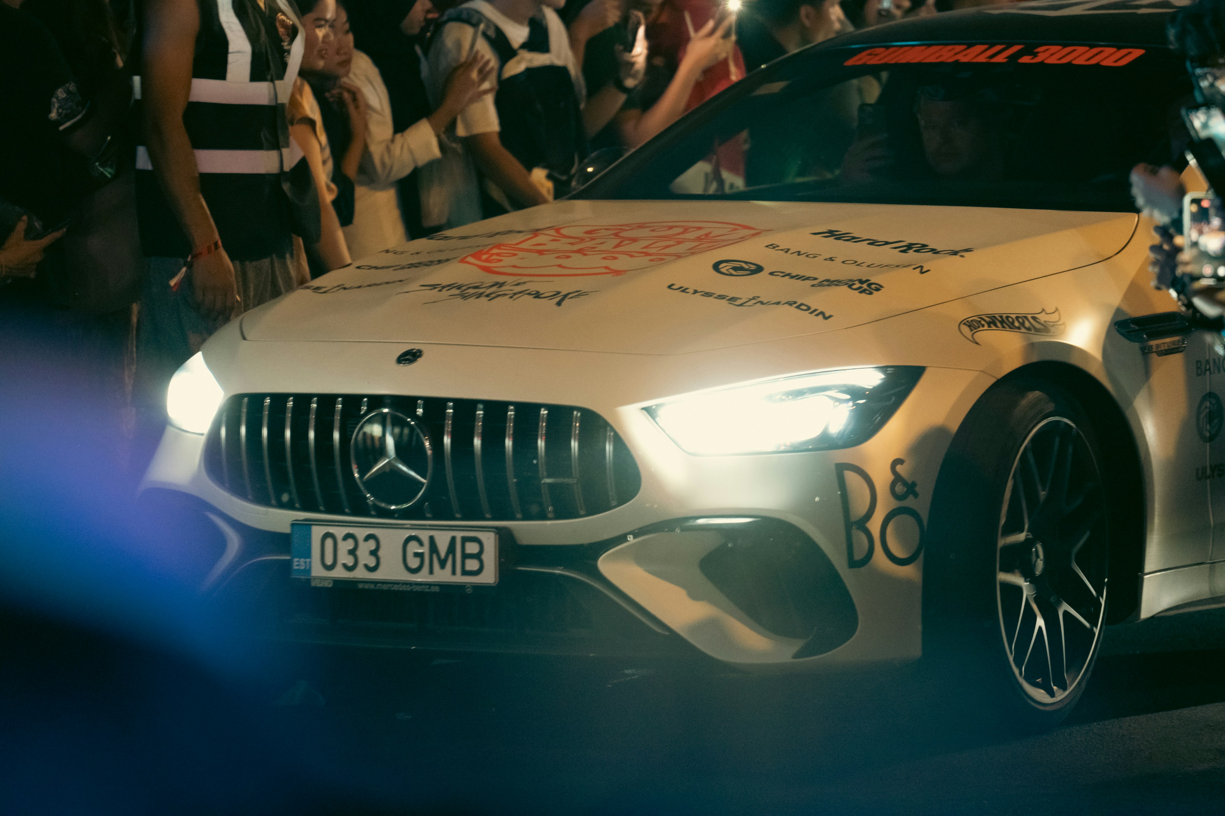 A white mercedes drives past a crowd of people.