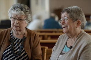 Two elderly women converse indoors, possibly at church.