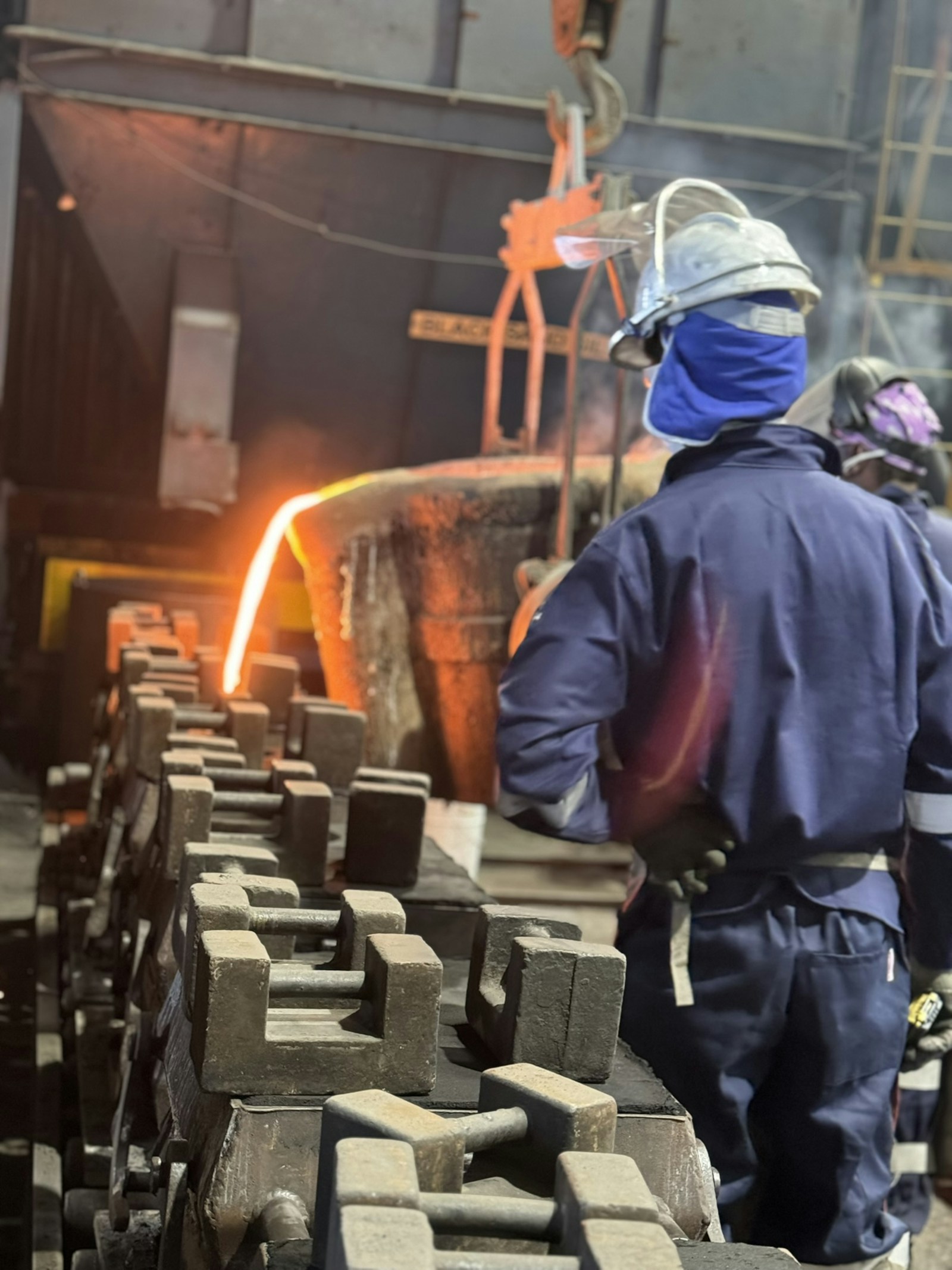 A foundry worker in a hard hat and protective jacket stands beside a bright stream of molten metal pouring into molds.