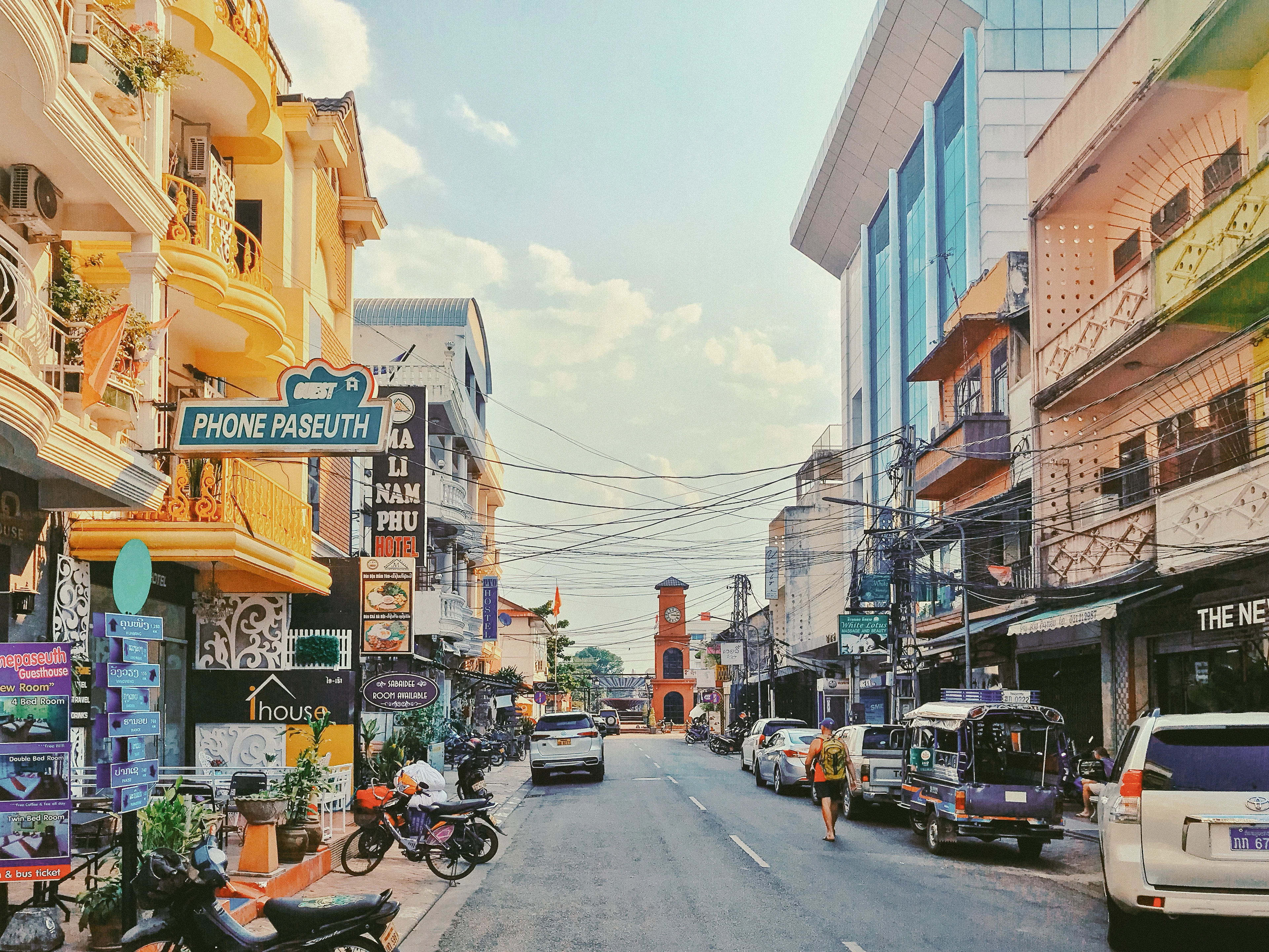 A guesthouse street in Vientiane's old quarter in late afternoon light, hotel and guesthouse signs stacked along balconied shophouses and the brick clock tower standing at the end of the road