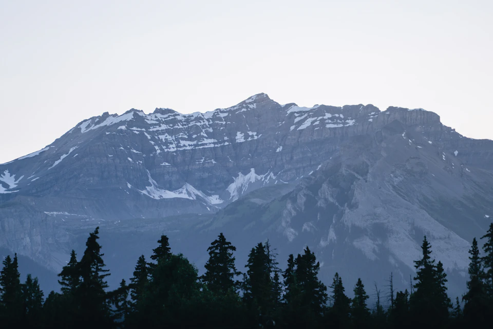 The image depicts a majestic mountain range with snow-capped peaks and a deep blue sky. The mountains, covered in snow, rise high into the sky, creating a stunning backdrop for the scene. The perspective is from below, looking up at the towering mountains that dominate the frame. In the foreground, there are trees scattered across the landscape, adding depth to the image and providing a sense of scale.