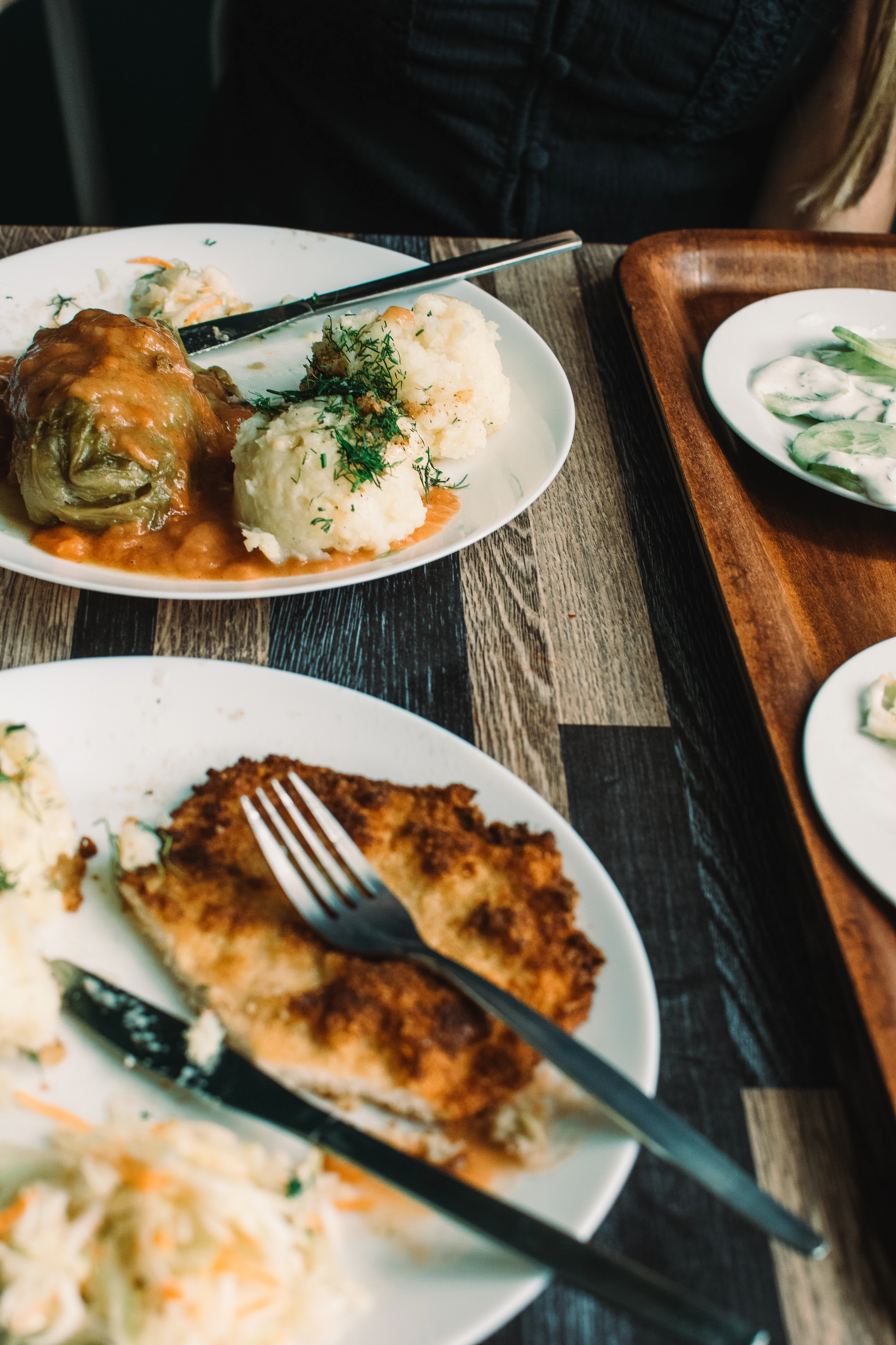 A milk-bar lunch on a dark wooden table in Warsaw: a cabbage roll in tomato sauce beside two scoops of dill-flecked mashed potato and a mound of white cabbage slaw, a breaded cutlet on the second plate with the cutlery resting on it, and a small dish of cucumbers in cream on a wooden tray alongside.
