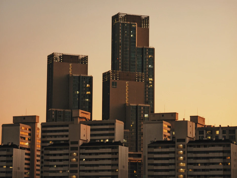 The image depicts a cityscape at sunset, with three tall buildings standing side by side on a street corner. The tallest building has a brown facade and is adorned with numerous windows that reflect the warm hues of the setting sun. The other two buildings have white facades and are topped with green roofs. The sky above them is painted in shades of orange and yellow, creating a stunning backdrop for the city's skyline.
