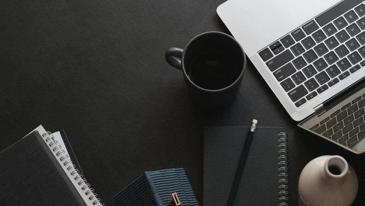 Laptop, buku catatan, dan gelas kopi di meja kerja gelap malam hari