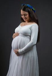 Pregnant woman wearing a white dress smiles gently.