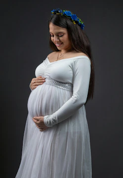 Pregnant woman wearing a white dress smiles gently.