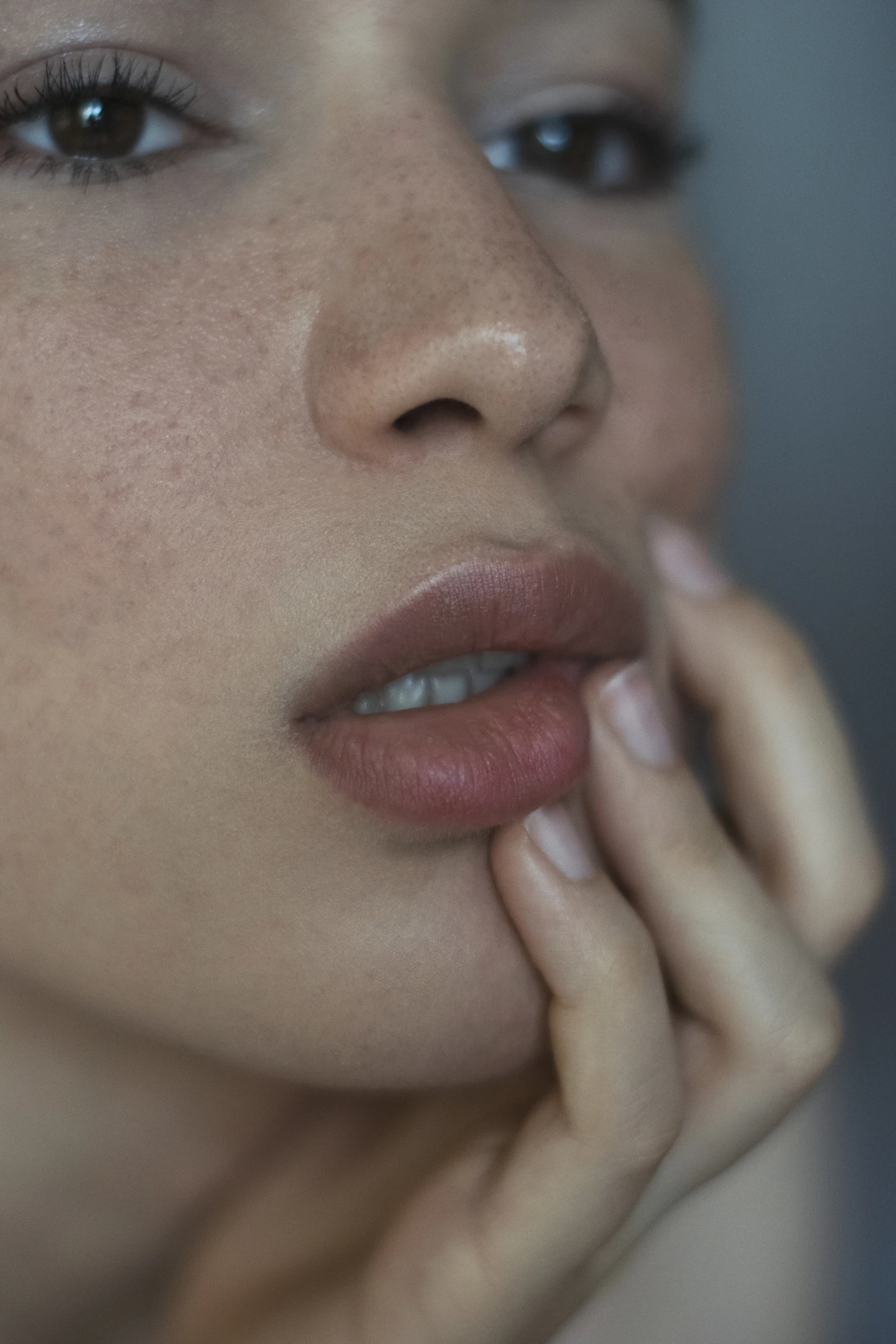 Soft close-up beauty portrait showing a woman’s eyes, nose and muted rosy lips, with her hand resting against her cheek.