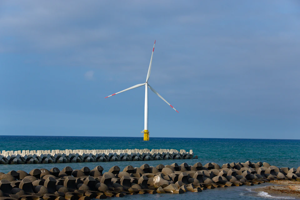 The image depicts a serene beach scene with a large white wind turbine standing tall on the right side, its blades facing towards the left side of the frame. The wind turbine is surrounded by a variety of rocks and boulders that add texture to the landscape. In the background, the vast expanse of the ocean stretches out, meeting the sky at the horizon line in the distance.