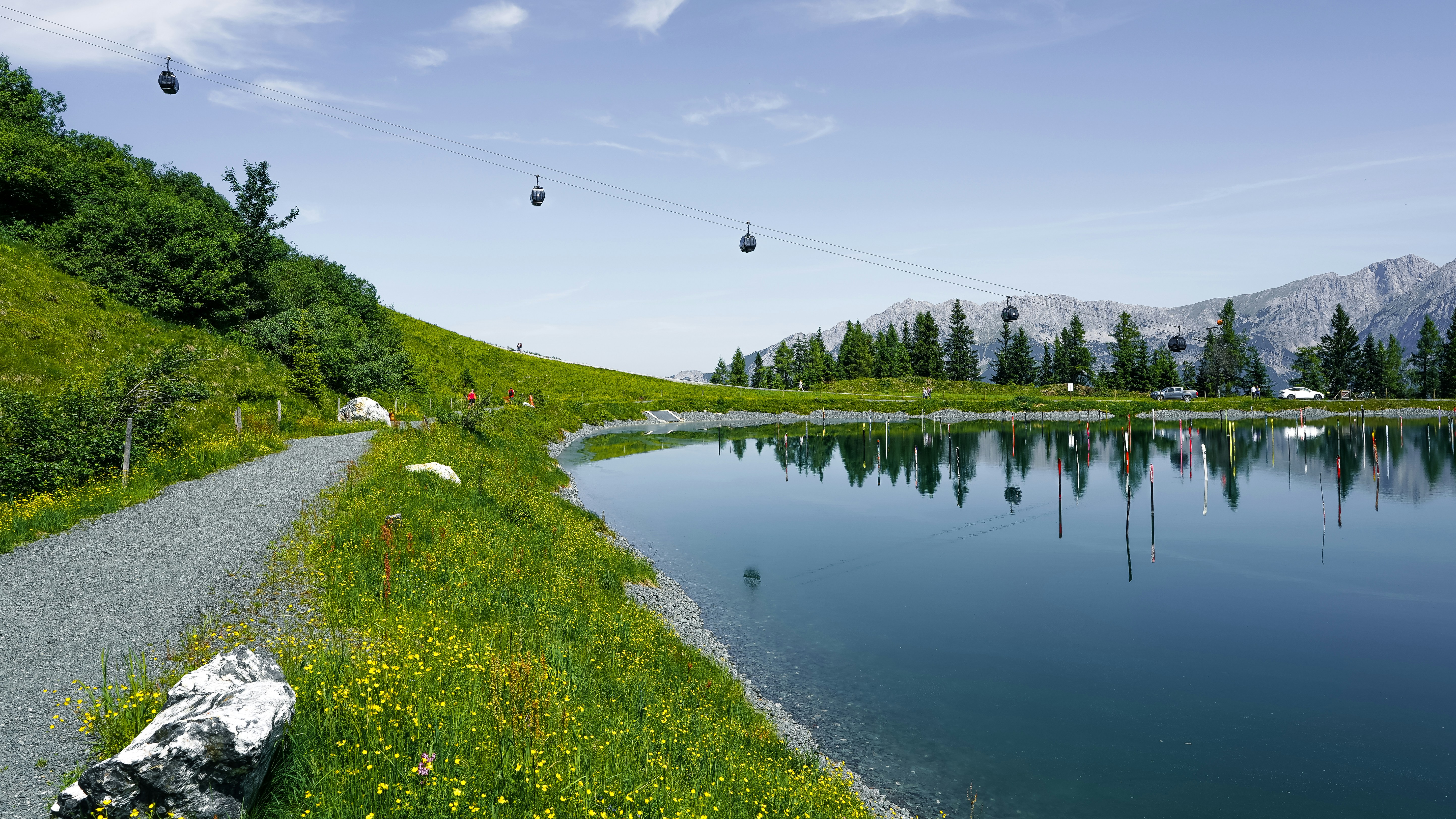 Scenic lake with a pathway, cable cars, and mountains.