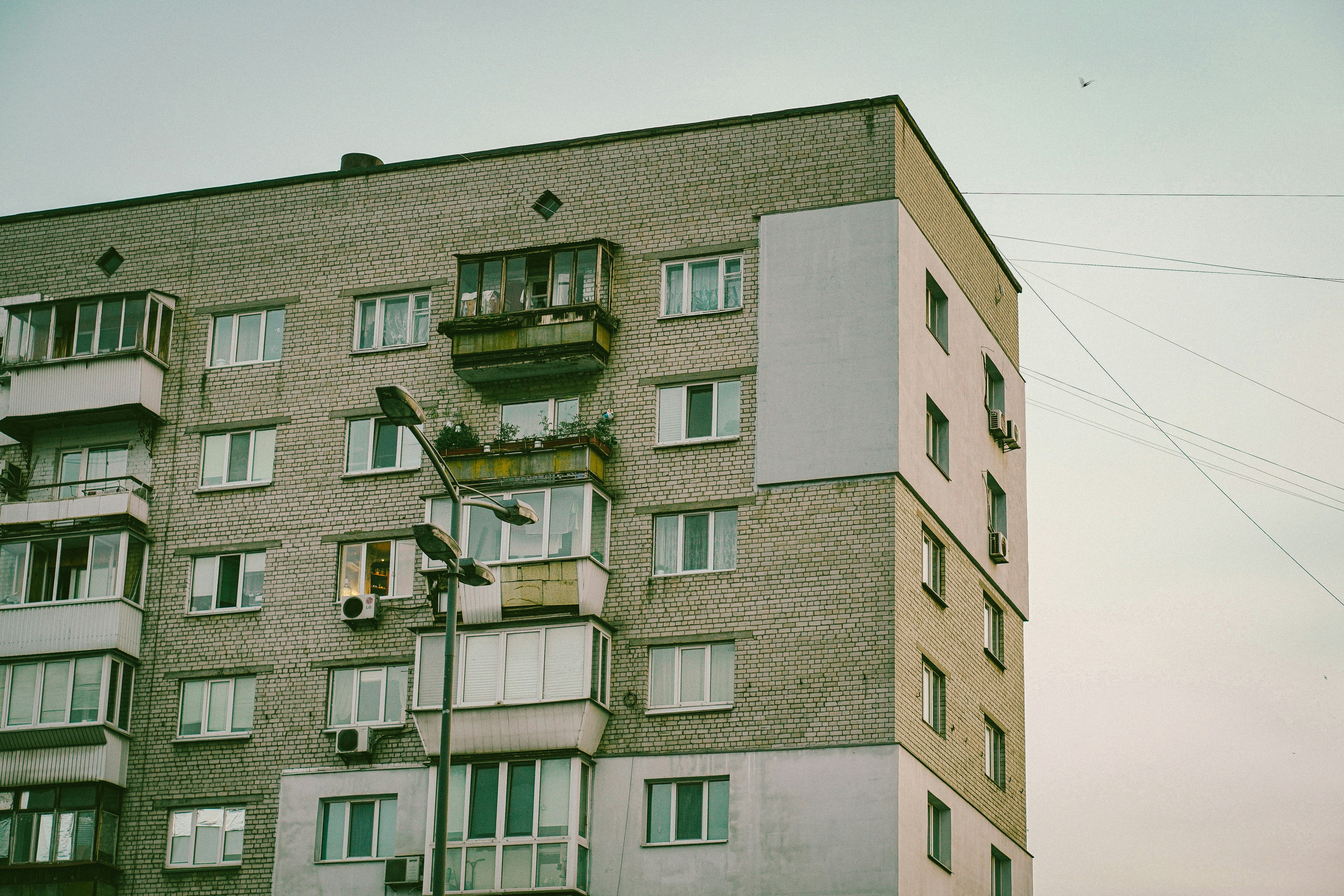 Muted grey brick apartment building with white windows and enclosed balconies, featuring a street lamp, under a pale, overcast sky.