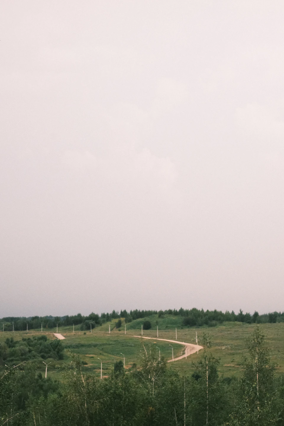 The image depicts a winding dirt road cutting through a lush, green landscape. The road is surrounded by an abundance of trees and shrubs on both sides, creating a sense of depth and perspective. The sky above is overcast with clouds, casting a soft light over the scene. The predominant colors in the image are shades of green from the vegetation and gray from the cloudy sky.