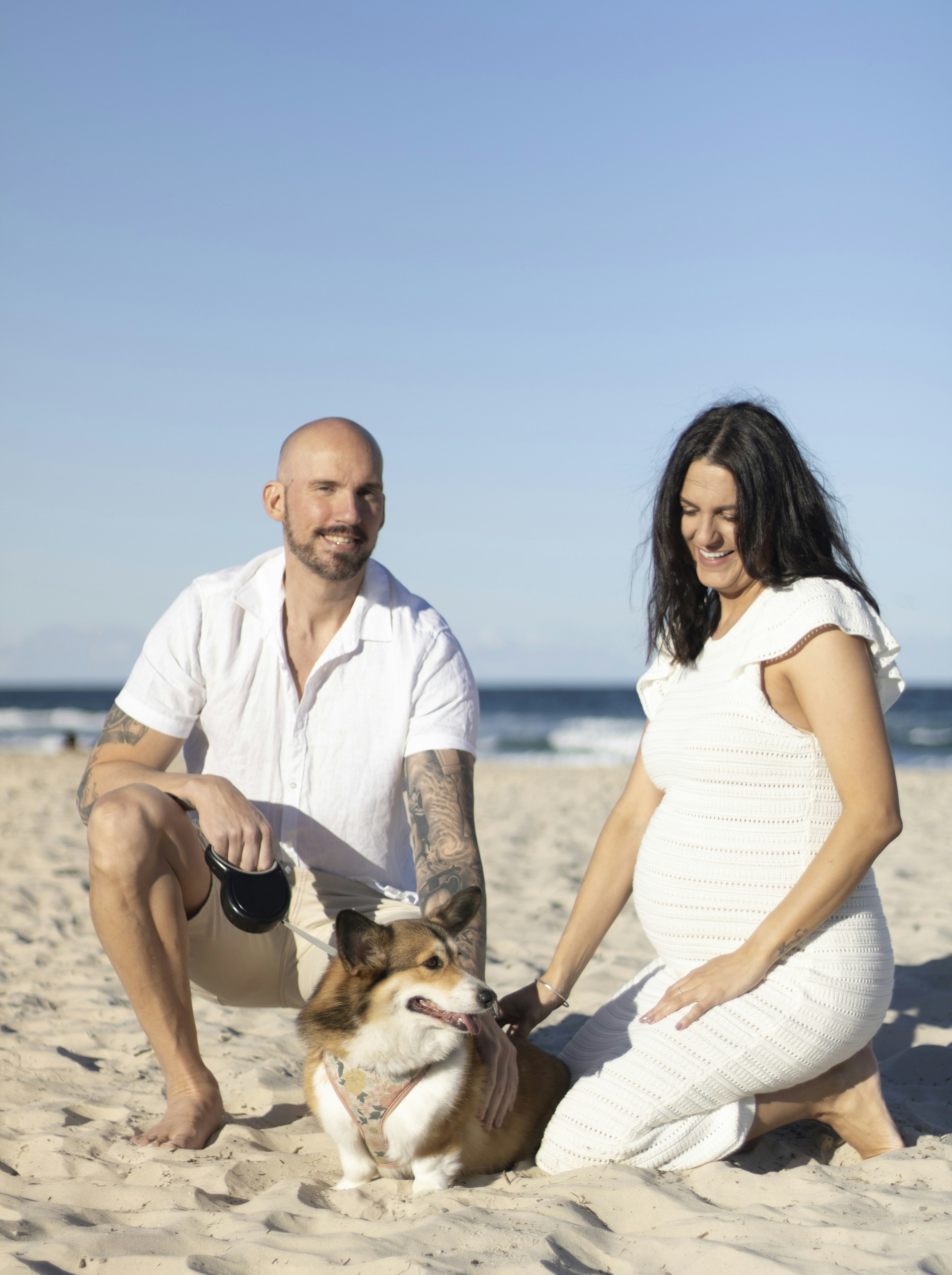 A pregnant couple smiles with their dog at the beach.
