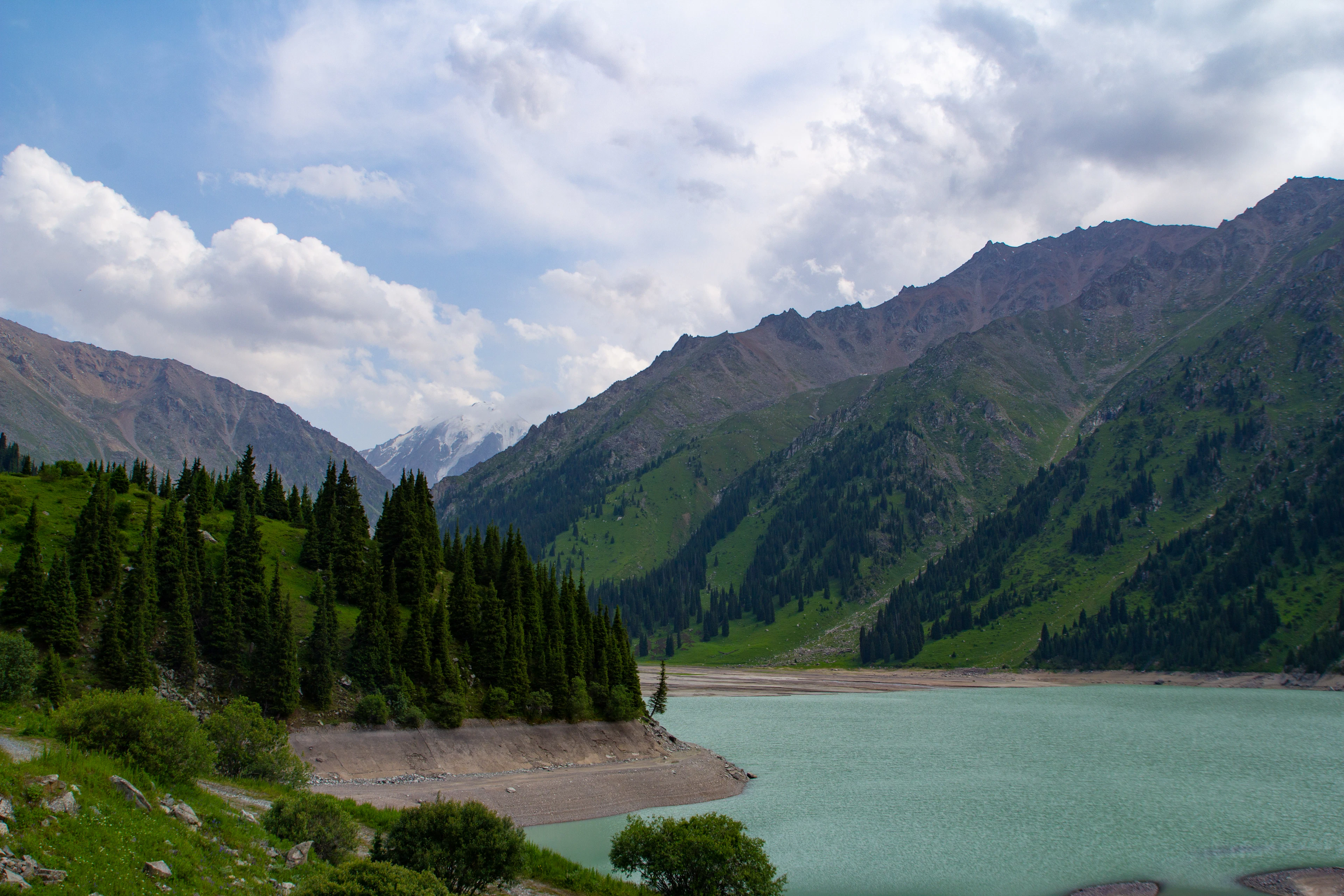 Big Almaty Lake's turquoise water stretches beneath forested slopes and jagged, partly snow-covered peaks.