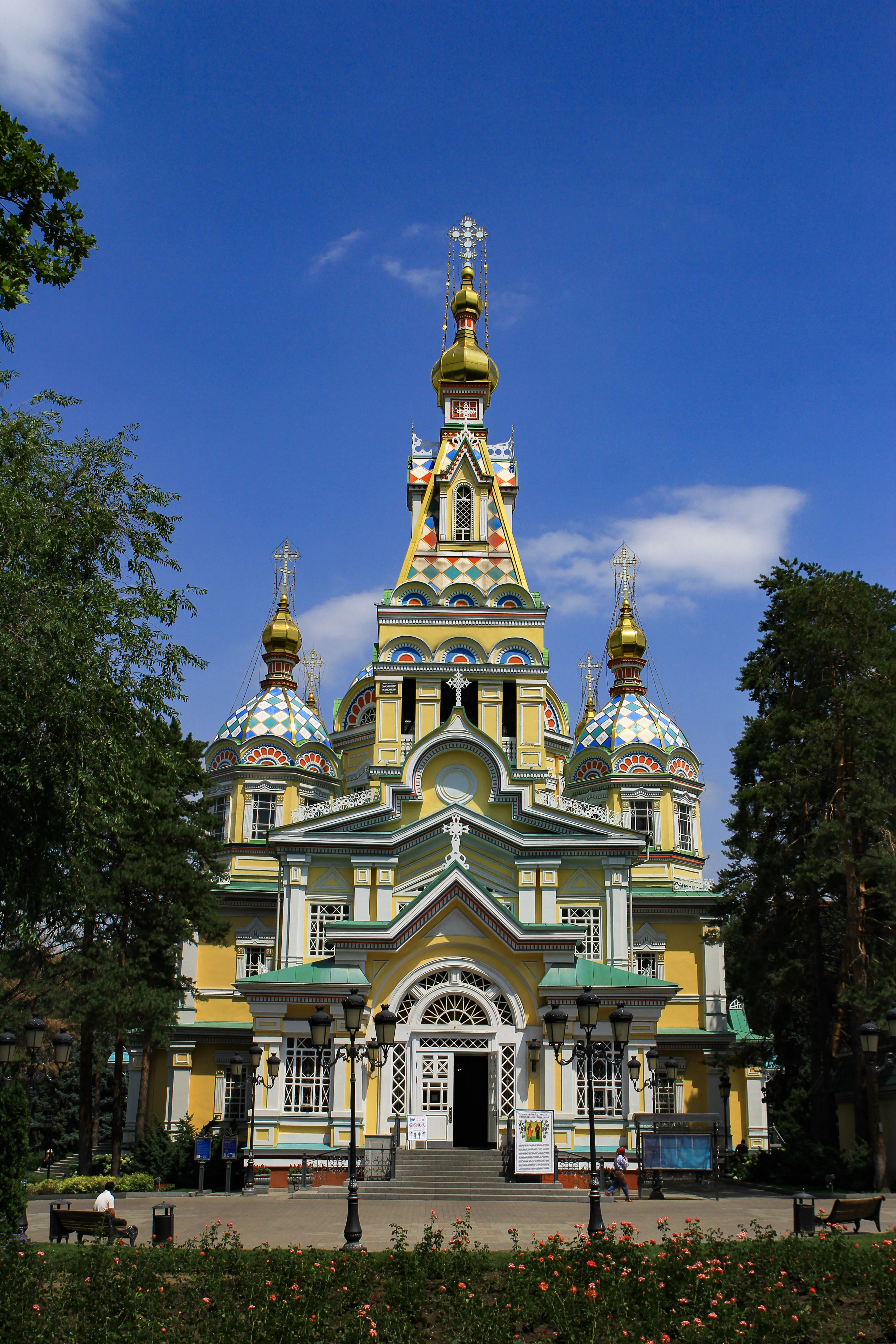 The wooden Ascension Cathedral in Almaty's Panfilov Park, its yellow facade, green porches and painted onion domes bright against a deep blue sky, with roses flowering in front