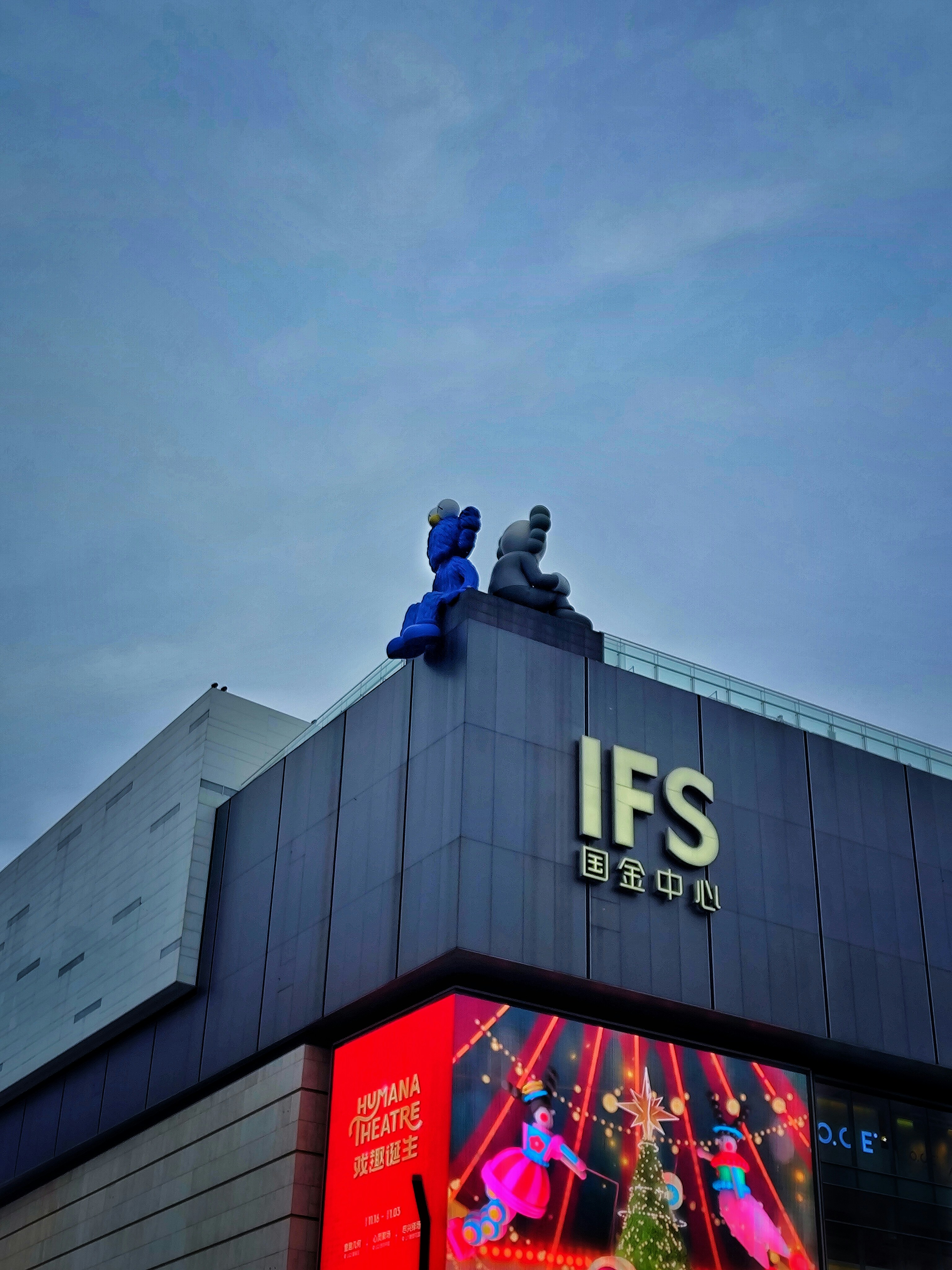 曇り空の下、建物の頂上にそびえるカウズの彫刻。