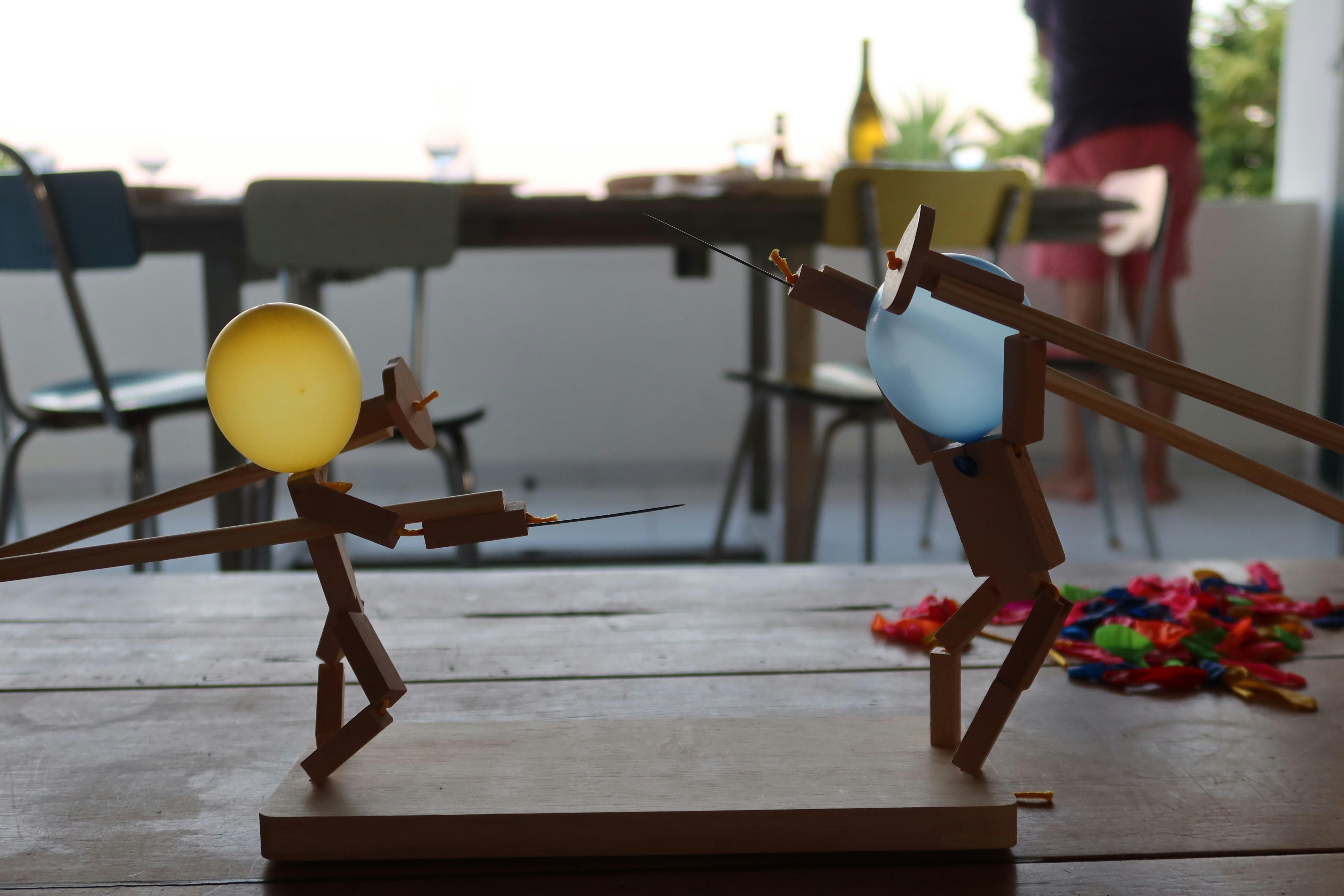 A playful tabletop battle unfolds between two wooden stick‑figure puppets with yellow and blue balloon heads, their skewering rods poised as colorful popped balloon fragments scatter across a rustic wooden table in a bright indoor‑outdoor dining space.