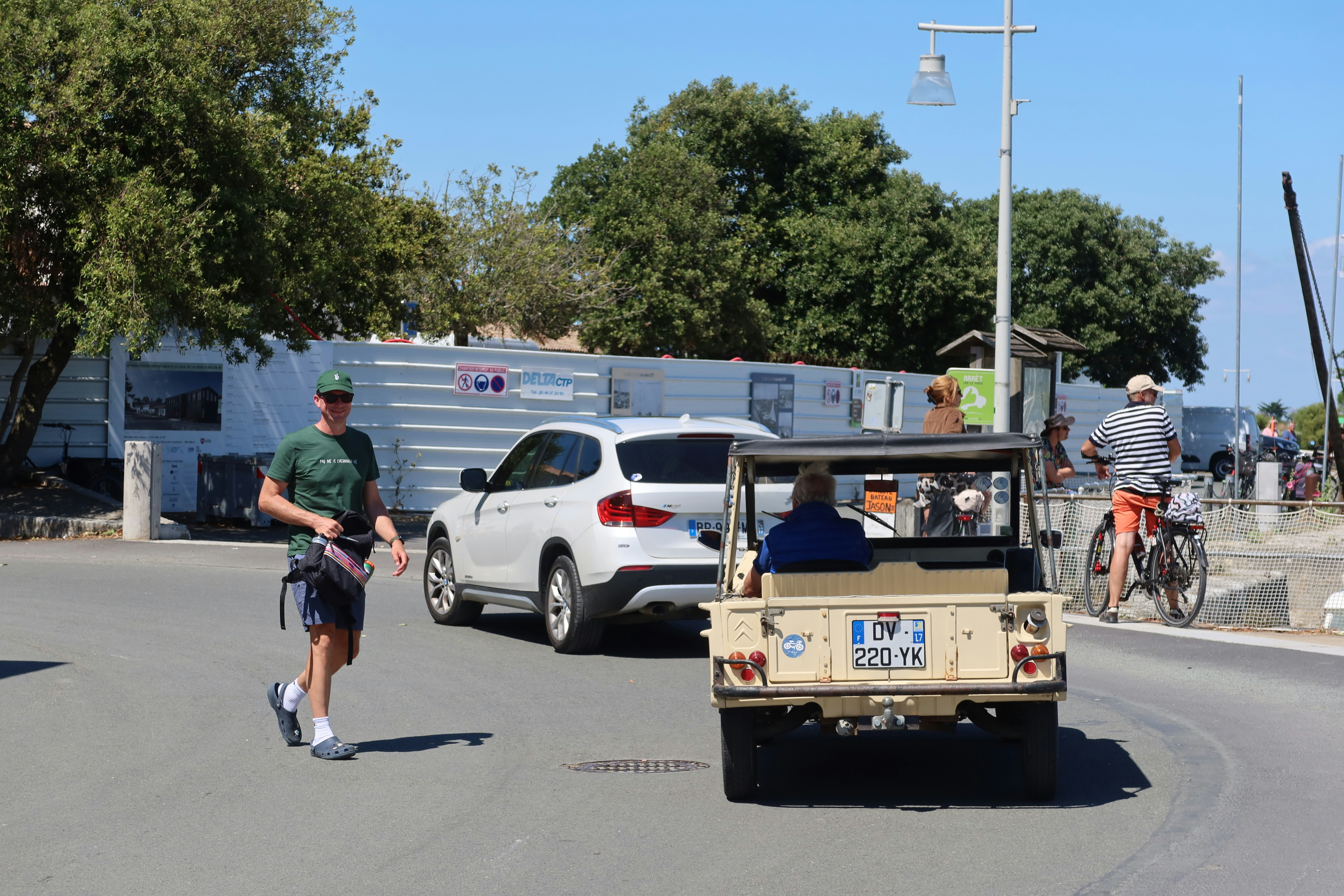A beige vintage Citroën Méhari with its top down rolls past a casually dressed man crossing a sunlit harbor street, as a cyclist and parked cars line the road under a clear blue sky.