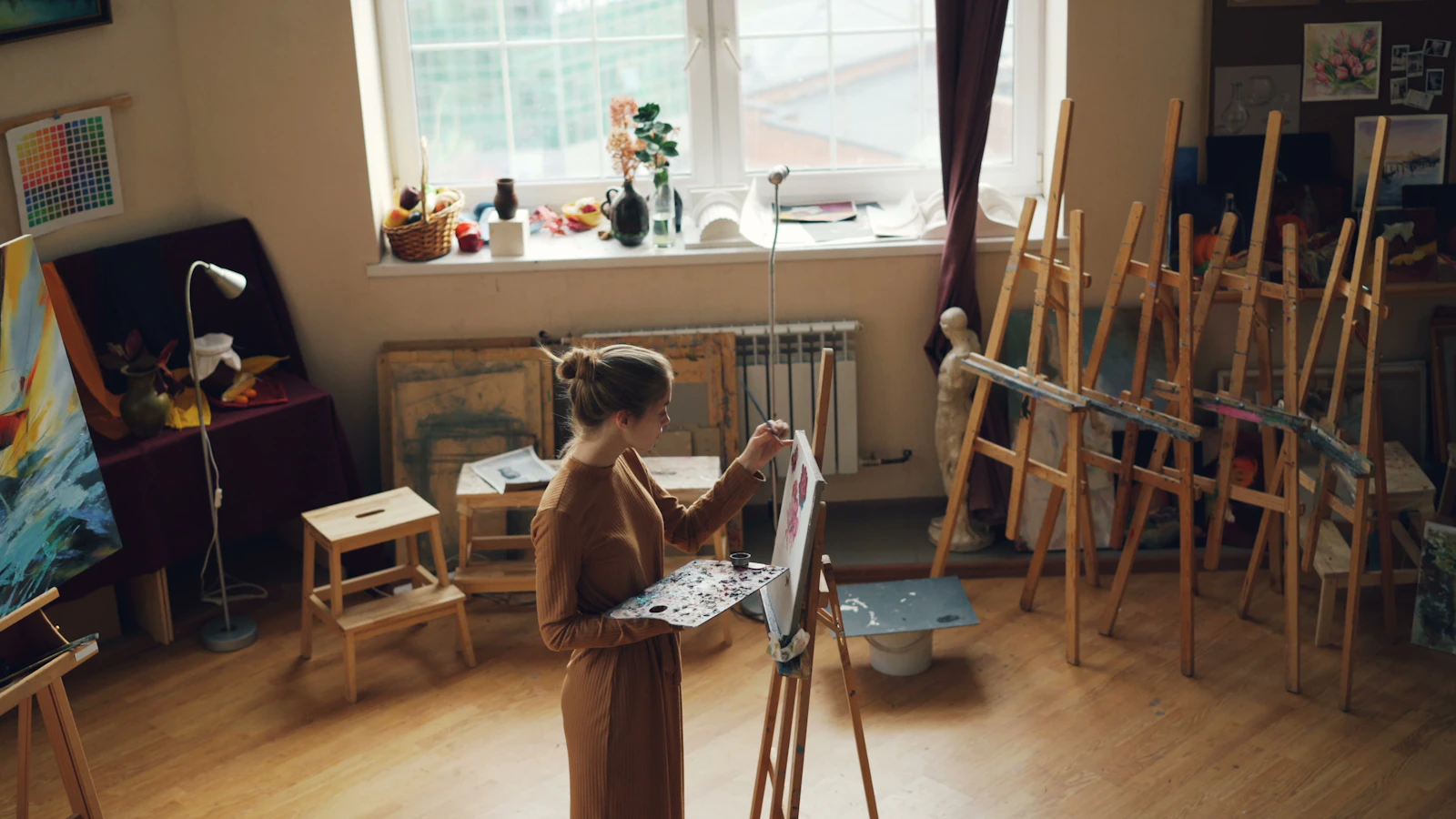An artist painting at an easel in a sunlit studio filled with canvases