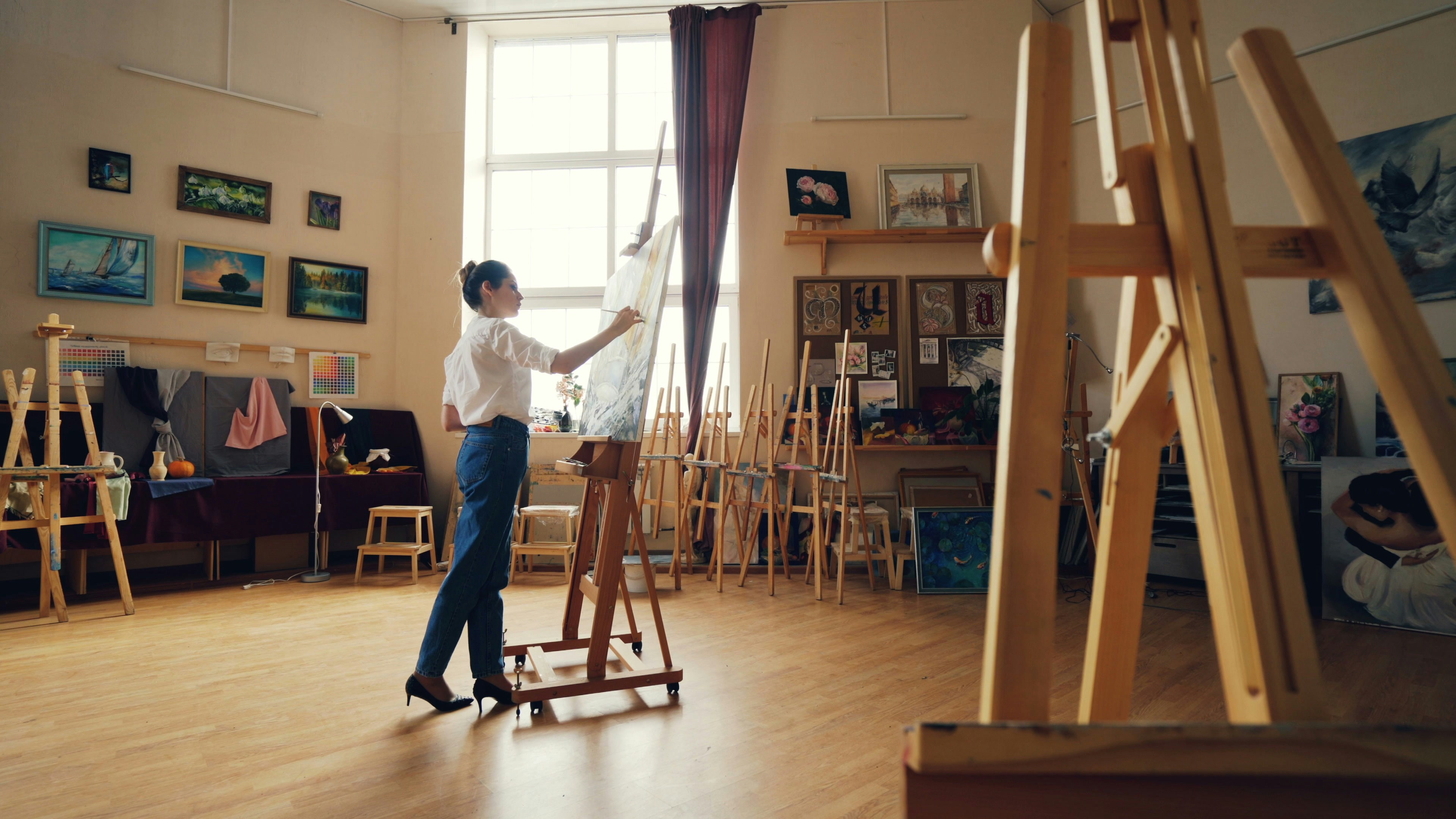 Serious young woman artist is working at picture painting with acrylic paints in arts school classroom with easels and artworks. Creative work, studio and people concept.