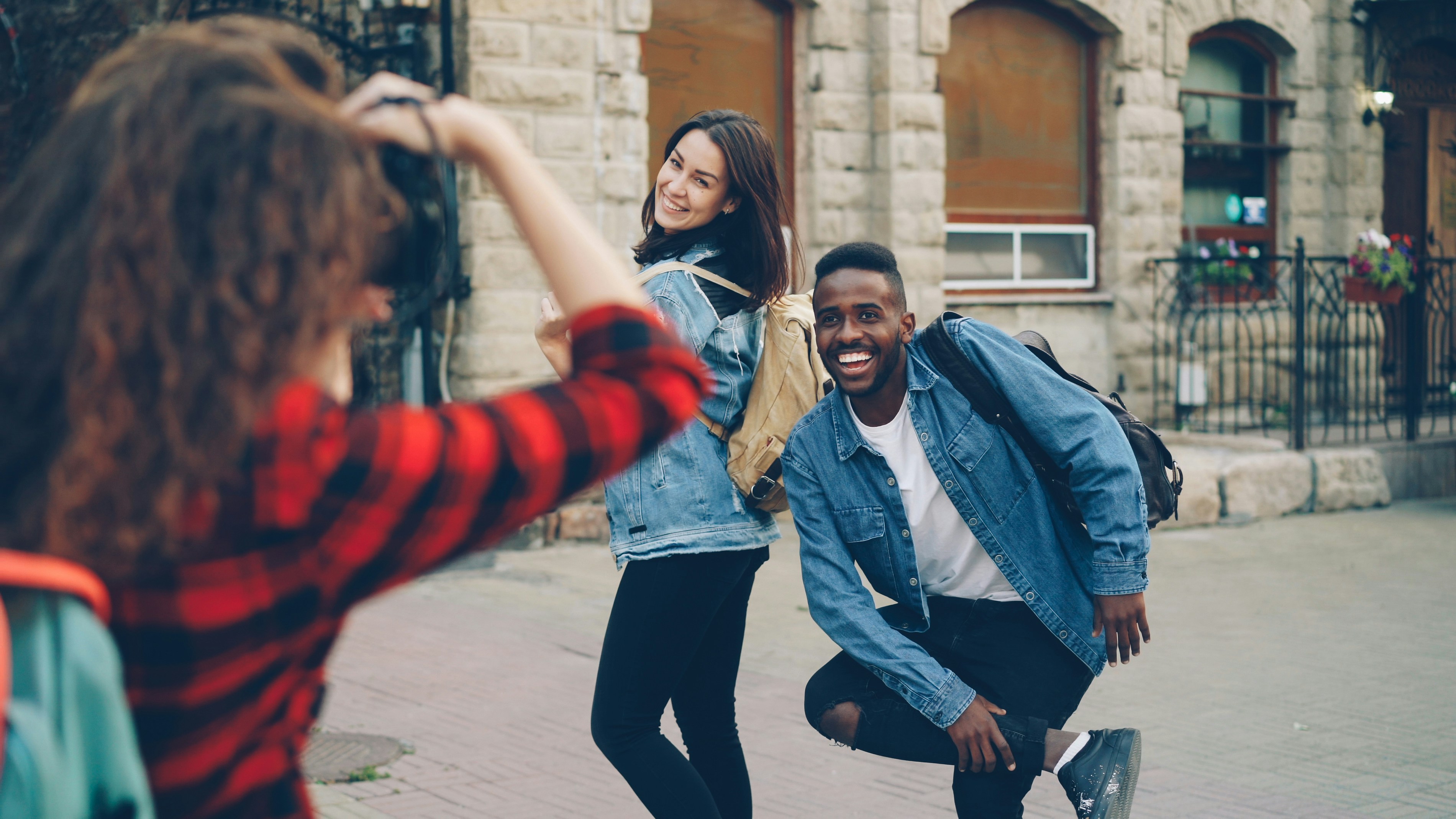 Creative girl and guy friends are posing for camera standing in the street in city while young woman with backpack is taking pictures. People are having fun and laughing.