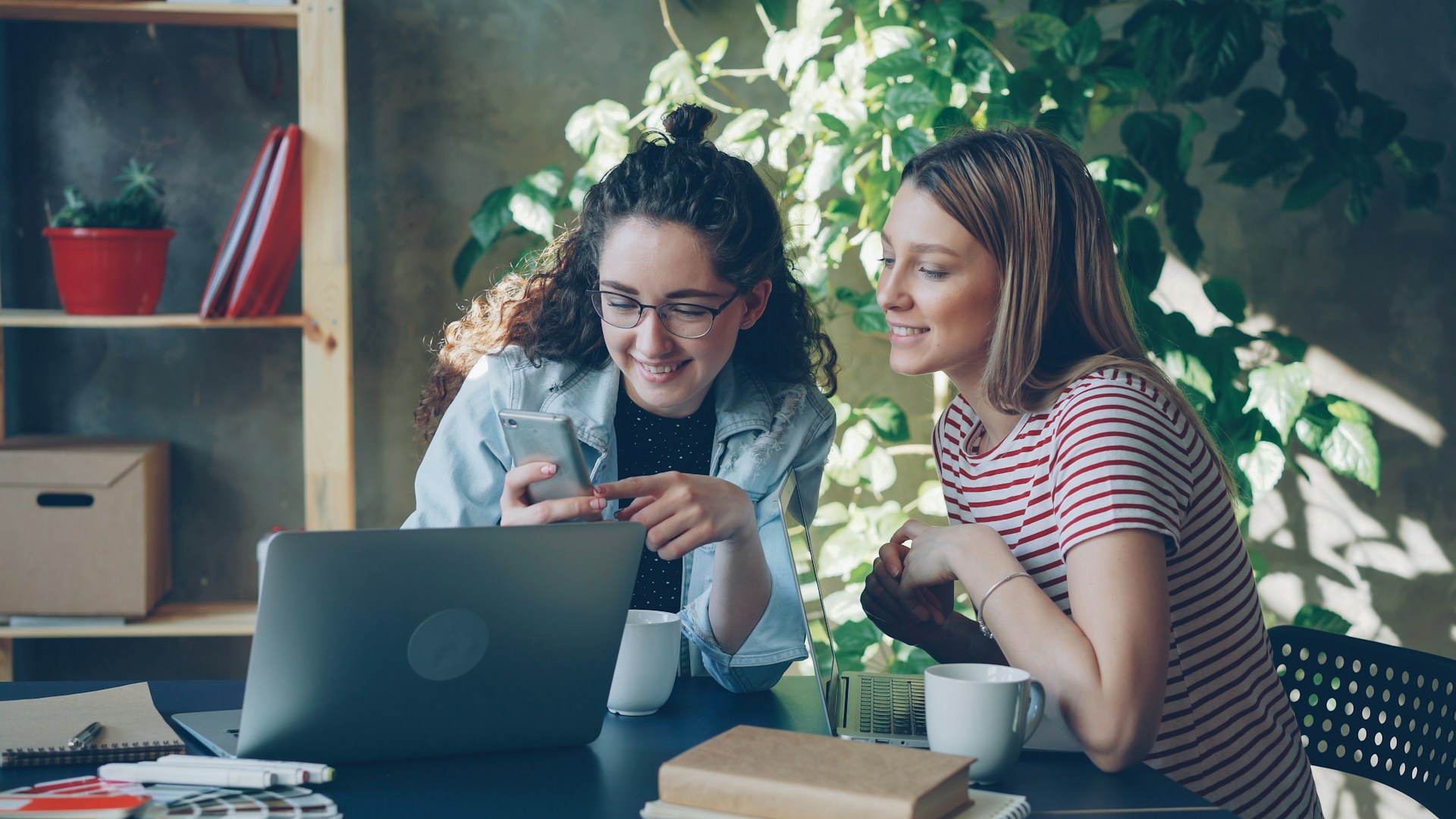Two women look at a laptop and smartphone.