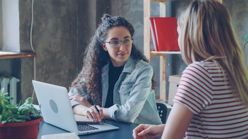 Two women are having a conversation at a desk.