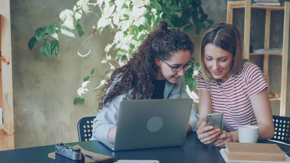 Two women look at a phone next to a laptop